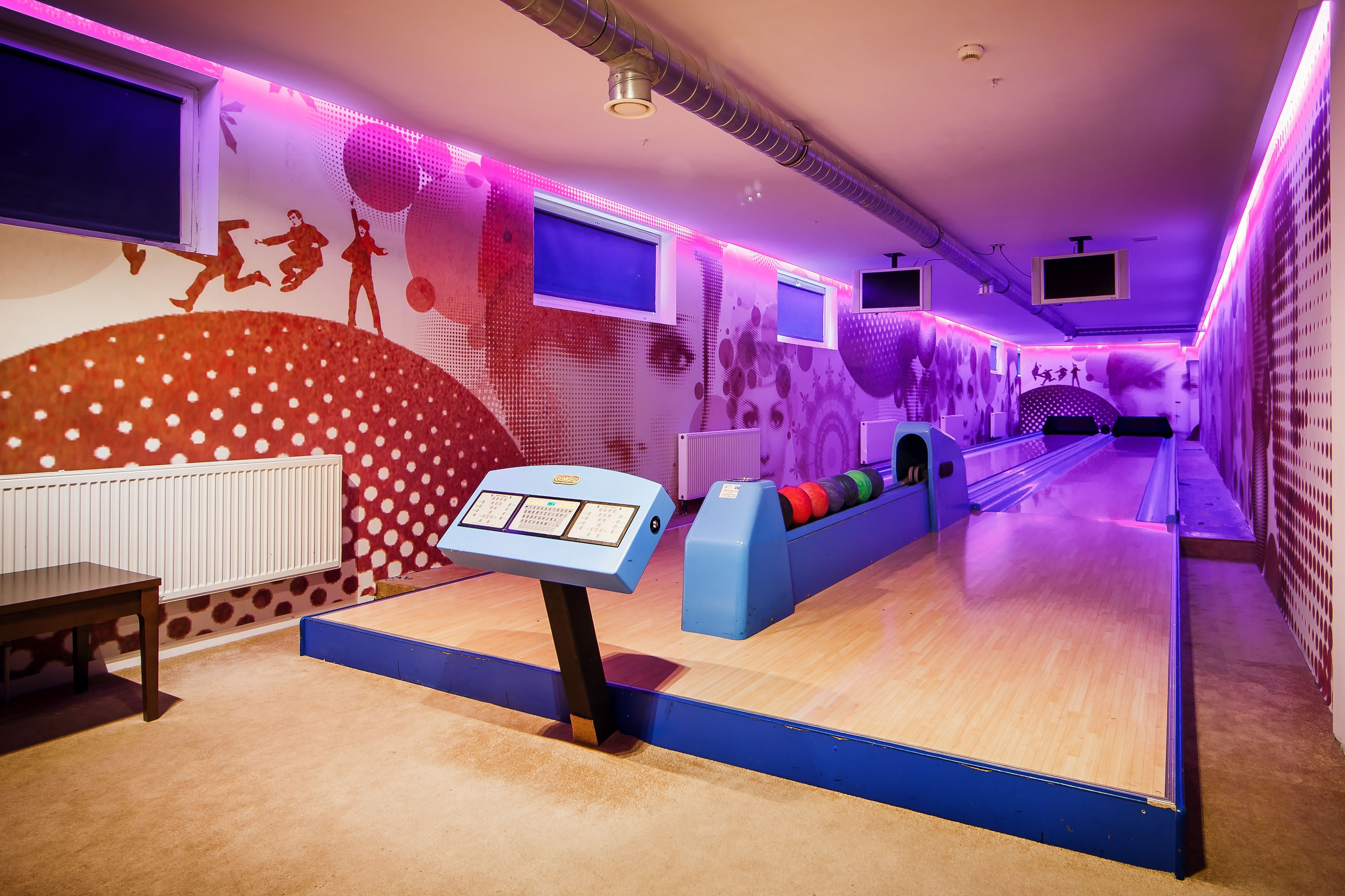 A bowling alley with a digital score display, multiple lanes, and a seating area with colorful cushions.