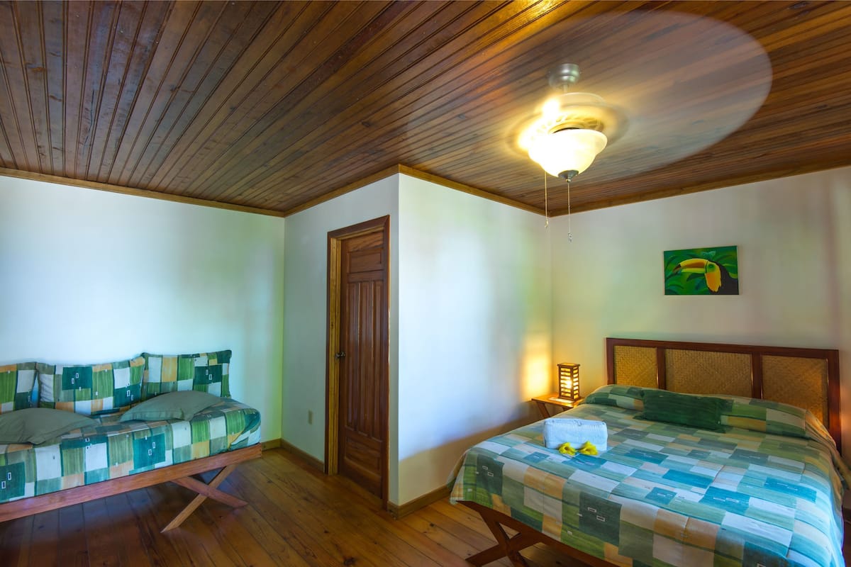 A room with a wooden ceiling, a bed with a quilt, a sofa with a patterned cover, and a door.