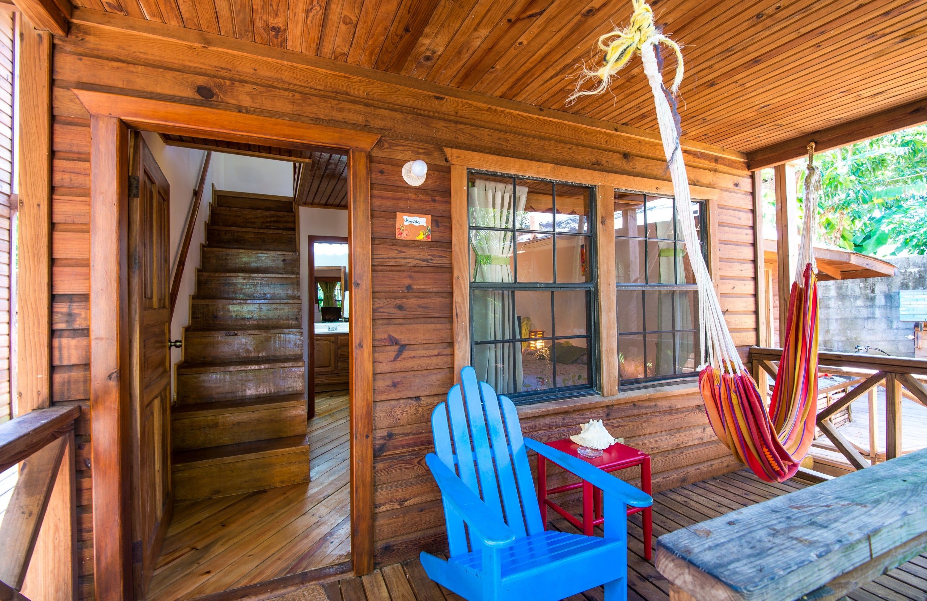 A wooden cabin with a wooden staircase leading to an upper level, a hammock, and a blue chair on the porch.