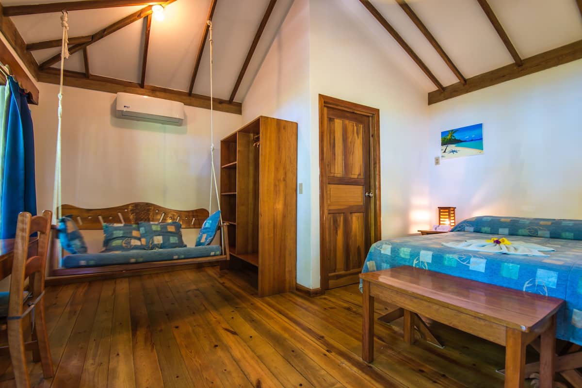 A room with a wooden ceiling, a bed with a blue and white checkered bedspread, a wooden bench, a wooden wardrobe, and a hanging light fixture.