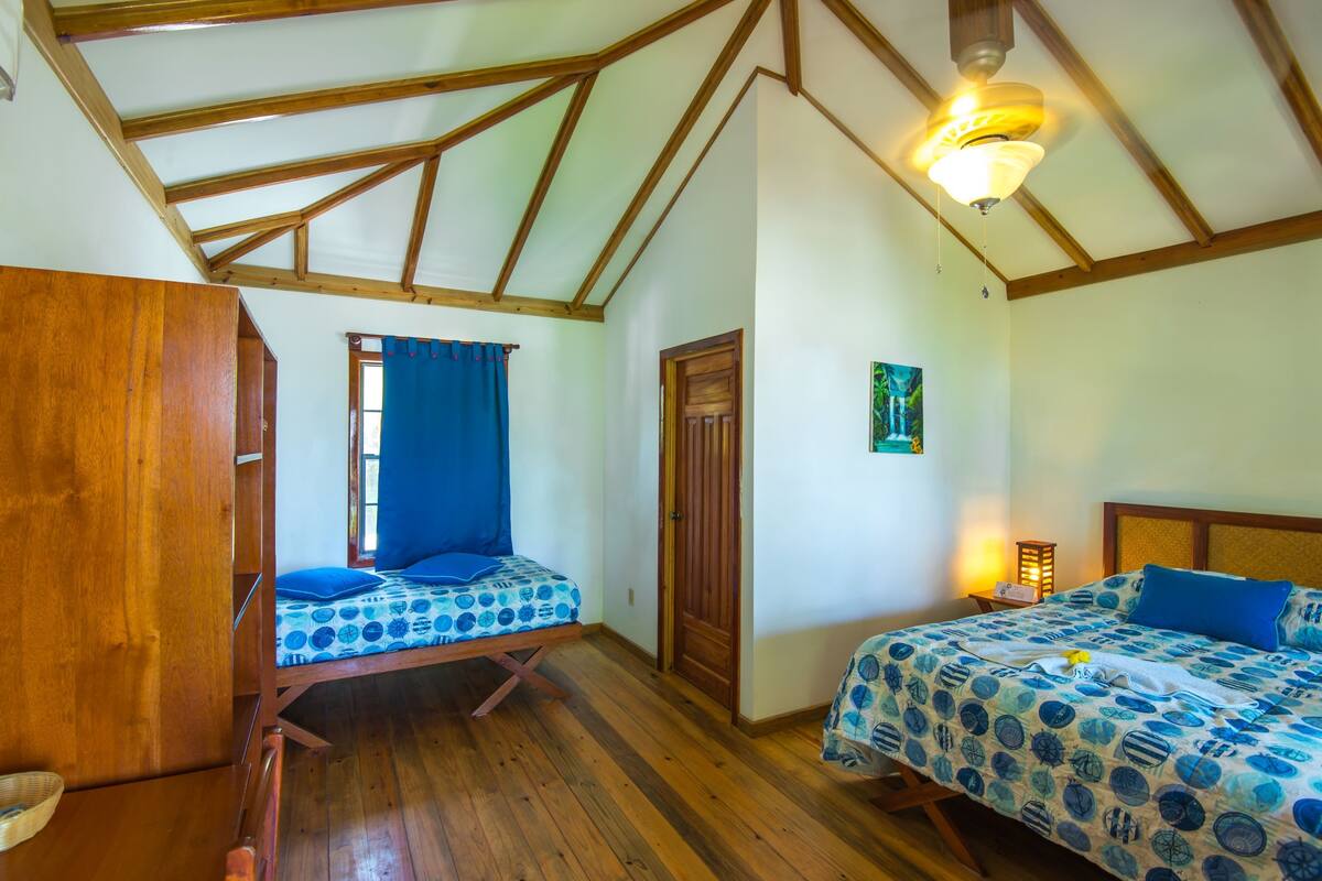 A room with two beds, wooden flooring, a ceiling fan, and a window with blue curtains.