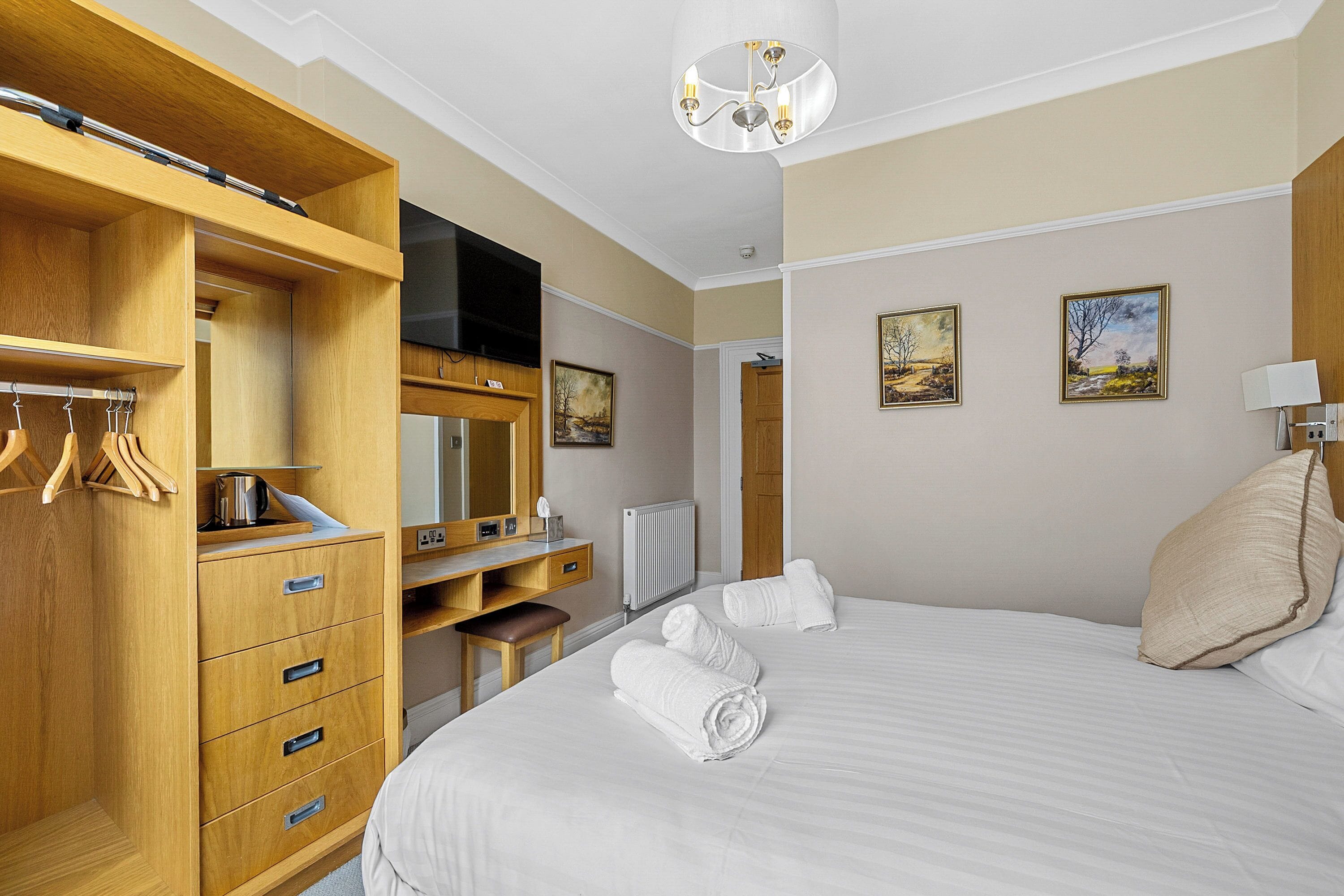A hotel room with a bed, a wooden wardrobe, a desk with a chair, a television, and two paintings on the wall.