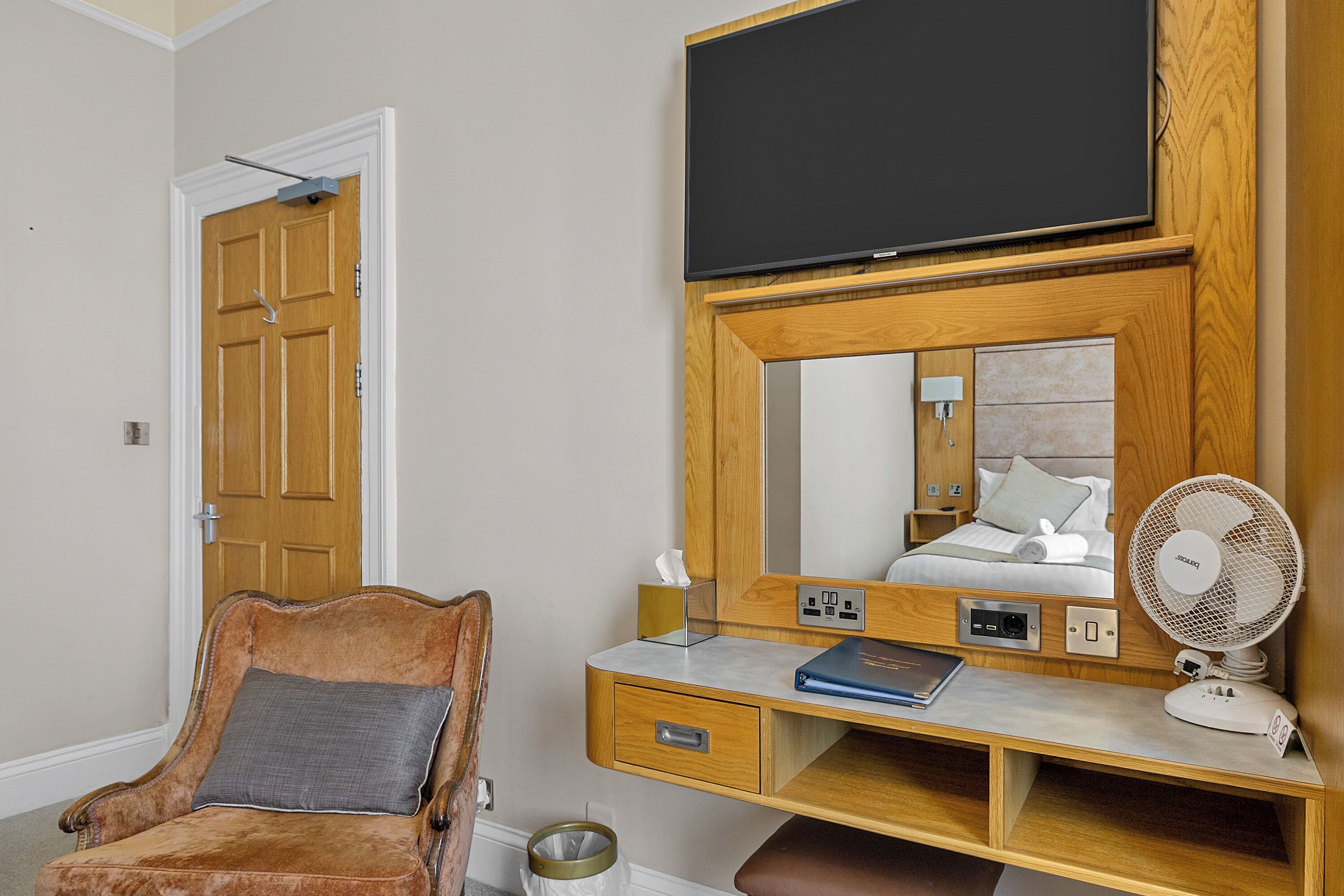 Une chambre d’hôtel avec un bureau en bois, un téléviseur fixé au mur au-dessus, une chaise et une table de chevet équipée d’un ventilateur.