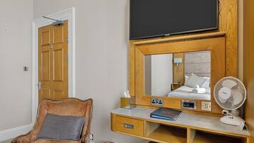 Une chambre d’hôtel avec un bureau en bois, un téléviseur fixé au mur au-dessus, une chaise et une table de chevet équipée d’un ventilateur.