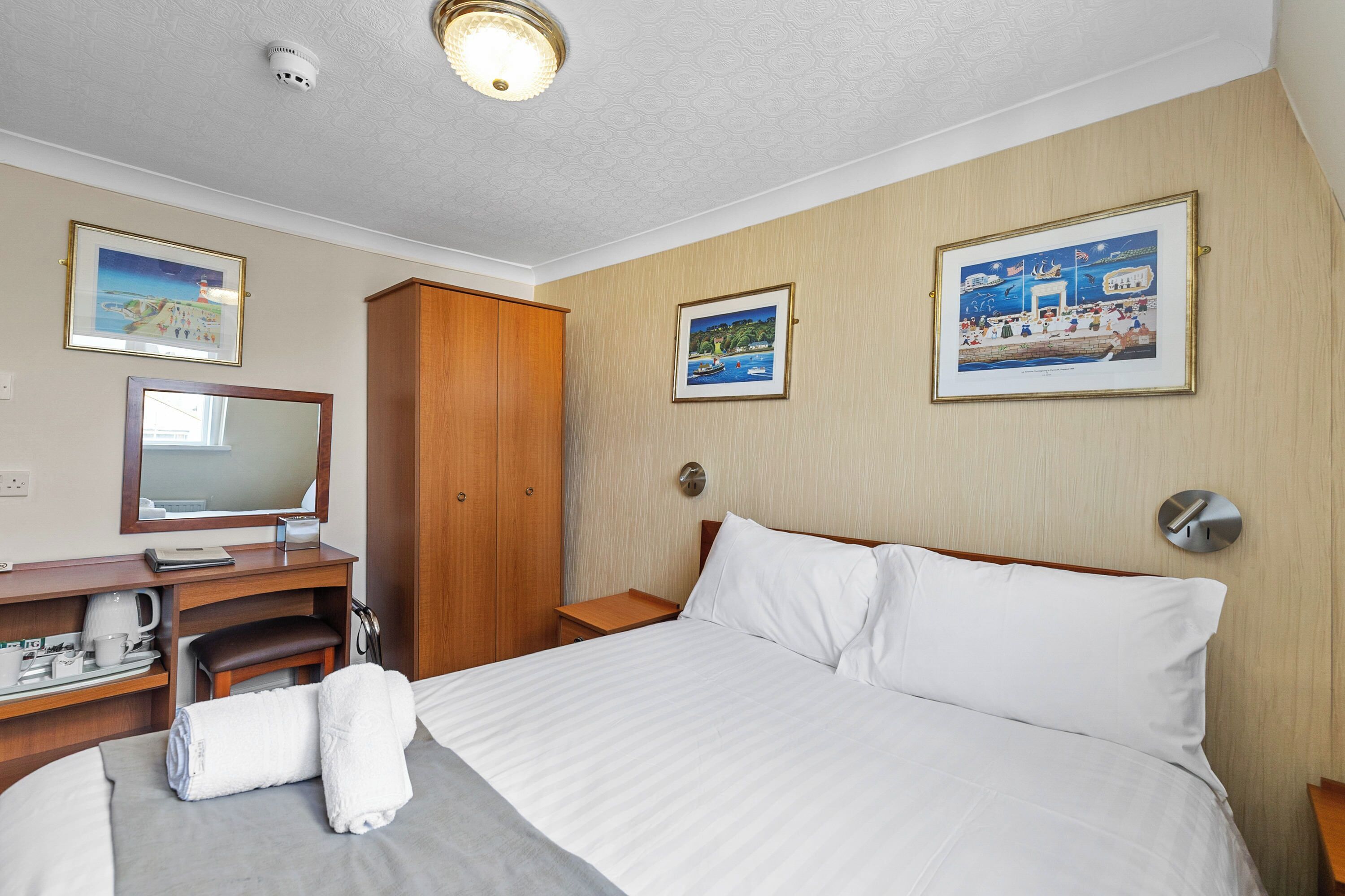 Habitación de hotel con cama, escritorio con espejo, cafetera, armario de madera y dos cuadros en la pared.