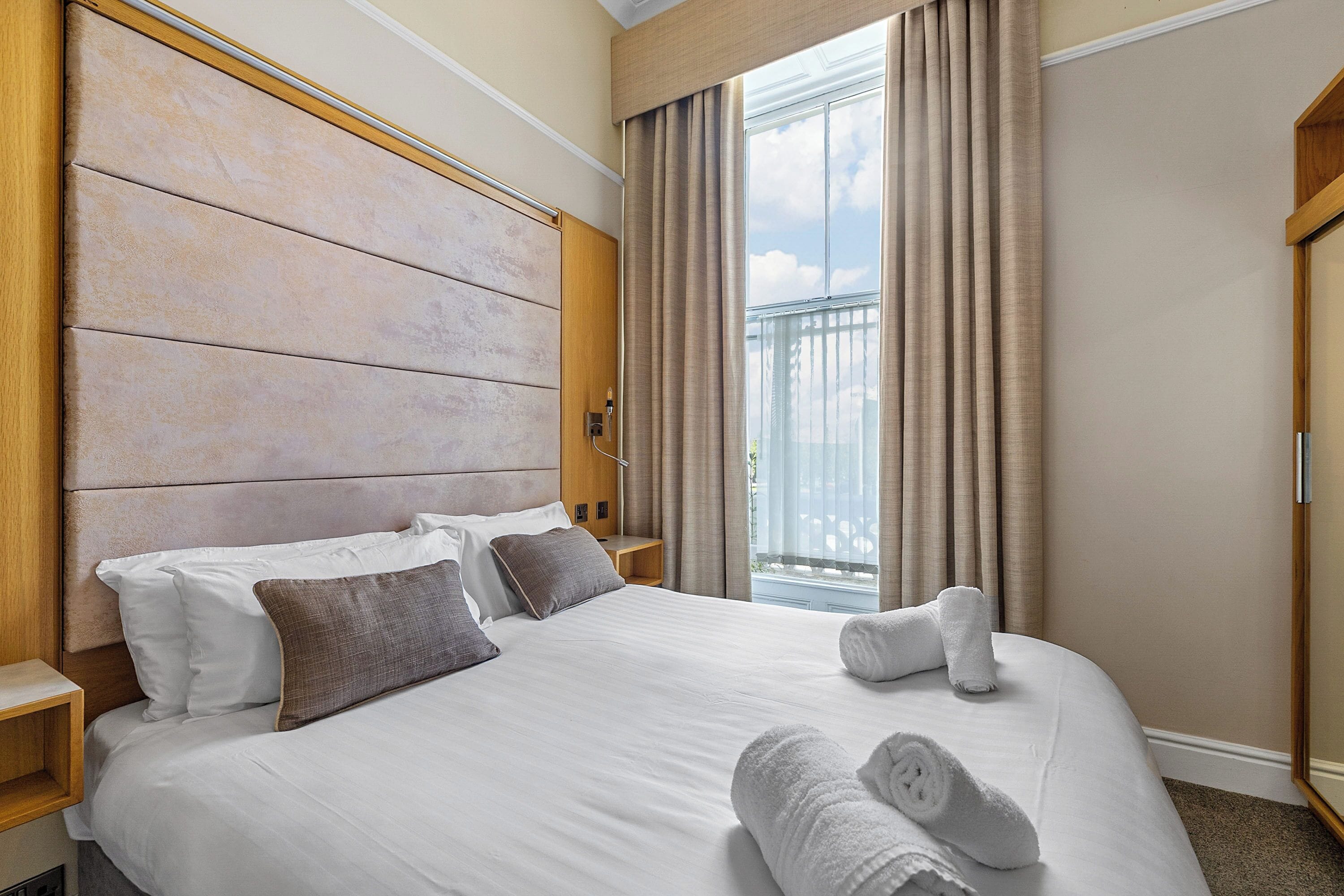 Quarto de hotel com uma cama grande, cabeceira de madeira, mesa de cabeceira e janela com cortinas.