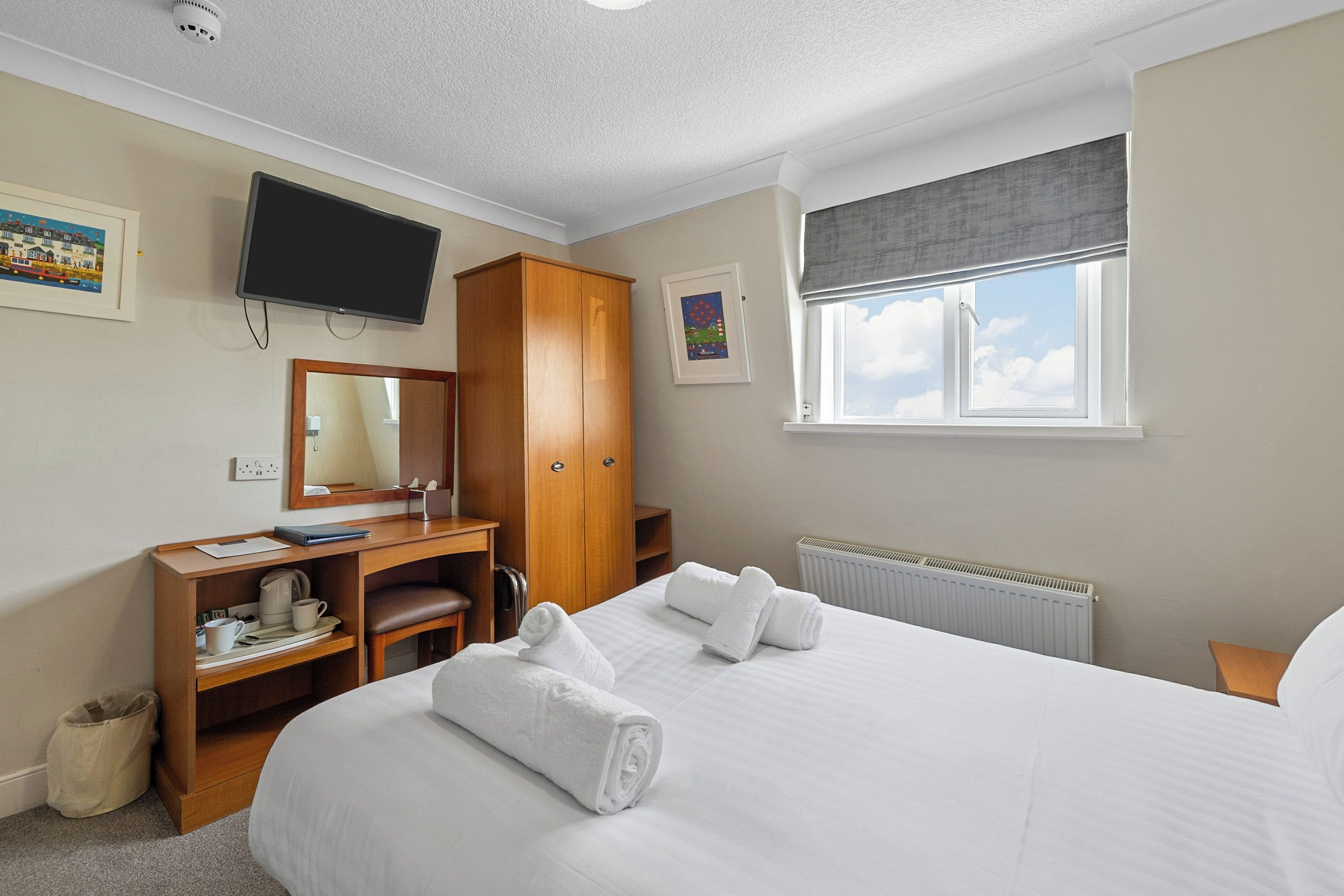 A hotel room with a bed, a desk with a chair, a television mounted on the wall, a window with a view of the sky, and a radiator.