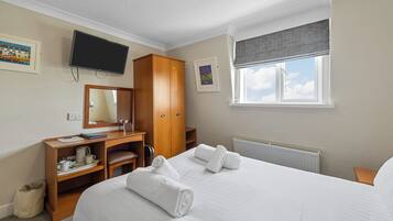 A hotel room with a bed, a desk with a chair, a television mounted on the wall, a window with a view of the sky, and a radiator.