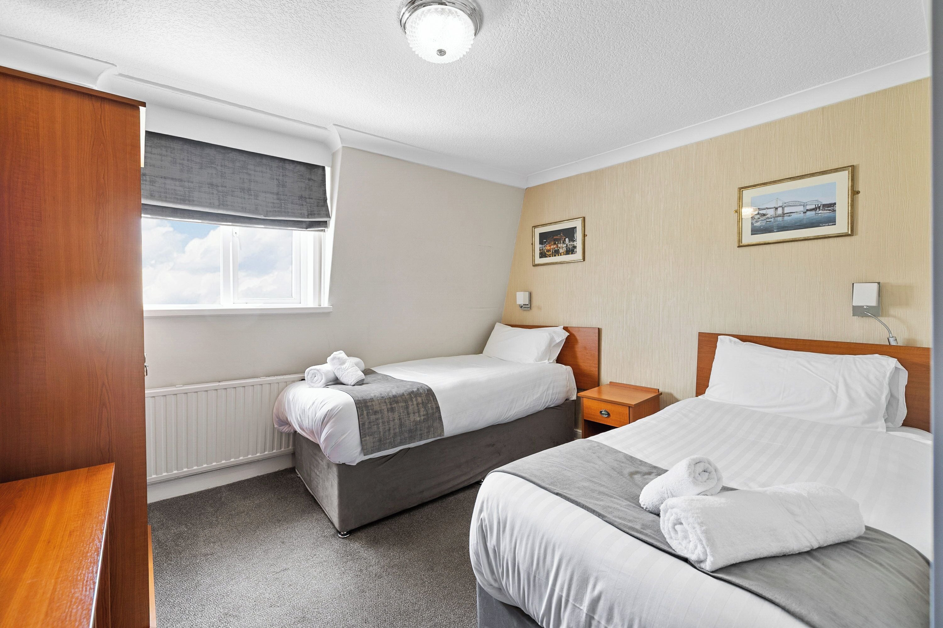 A hotel room with two beds, a wooden wardrobe, a window with a view, and a picture on the wall.