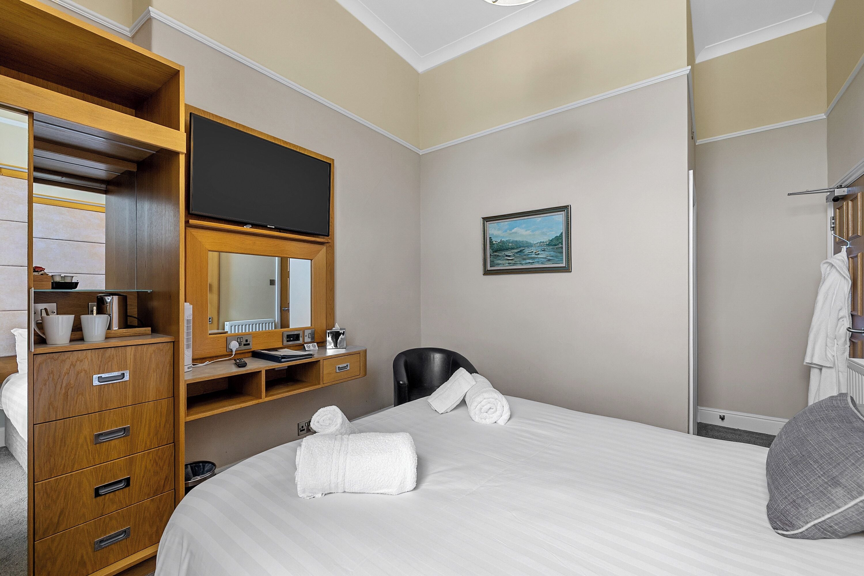 Quarto de hotel com cama, cómoda de madeira, televisão, pintura e casa de banho com porta-toalhas.