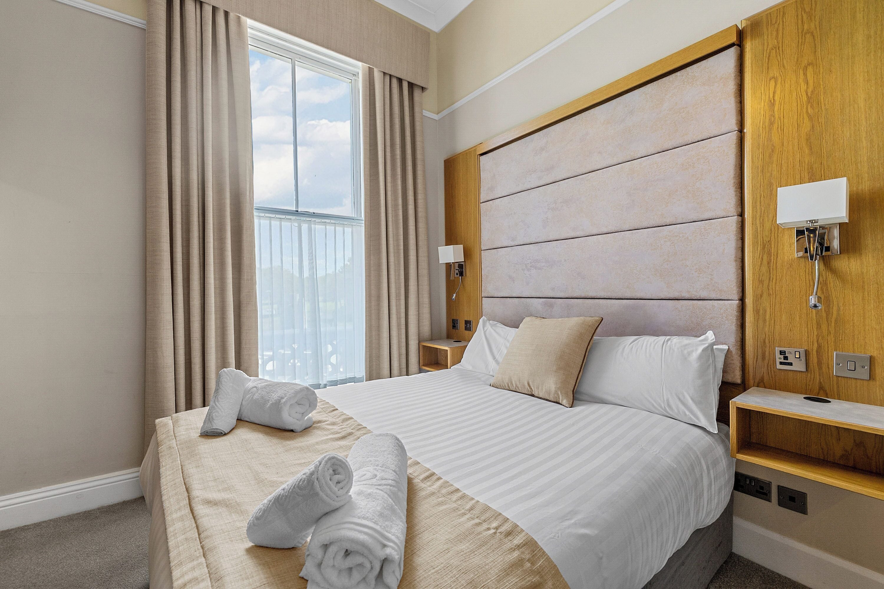 Habitación de hotel con una cama grande, dos mesitas de noche, un cabecero y una ventana con cortinas.