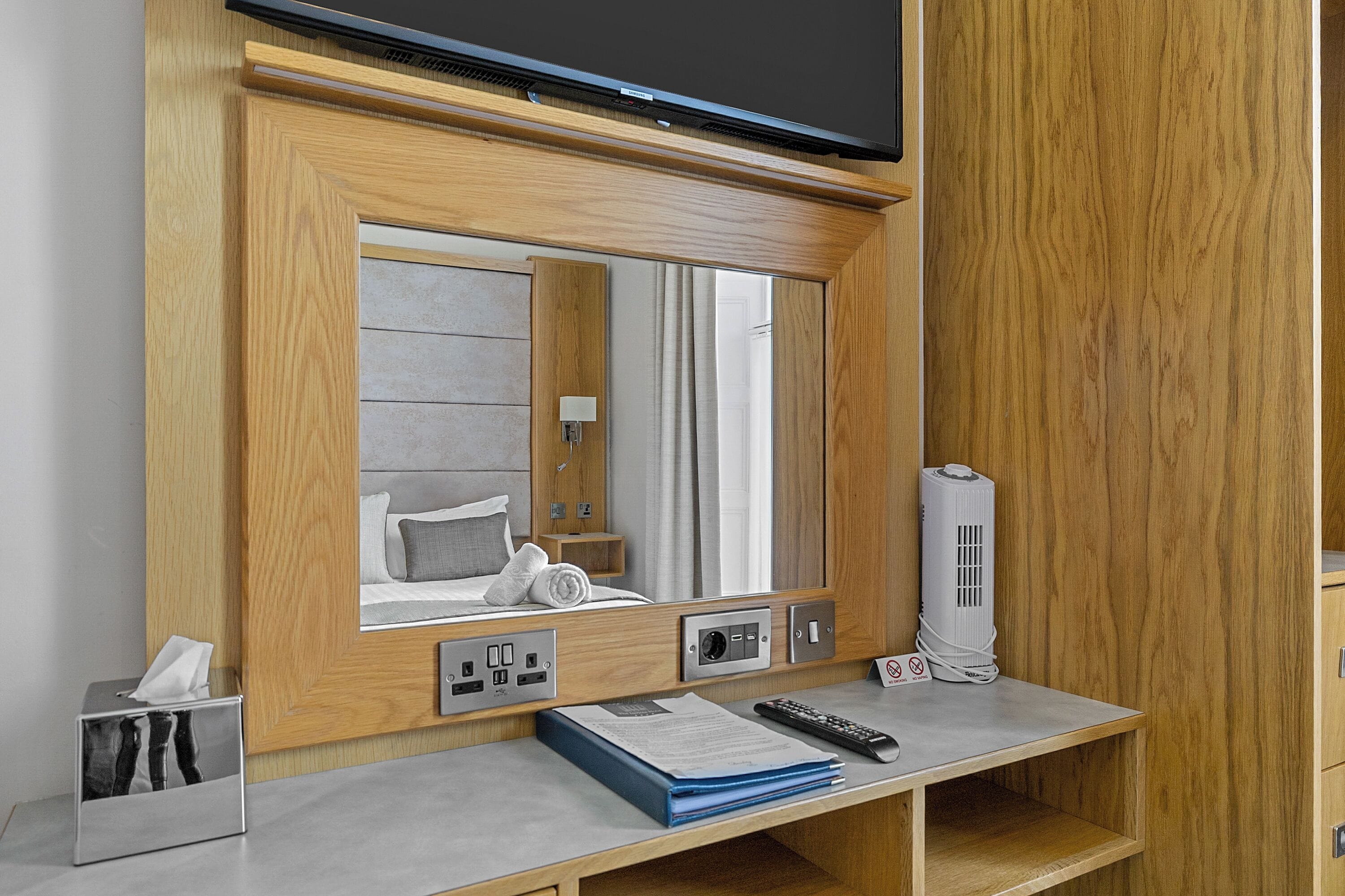 Hotelkamer met een spiegel met houten lijst, een bed met kussens, een nachtkastje, een lamp, een verwarming, een bureau met een afstandsbediening, een zakdoekendoos en een boek.
