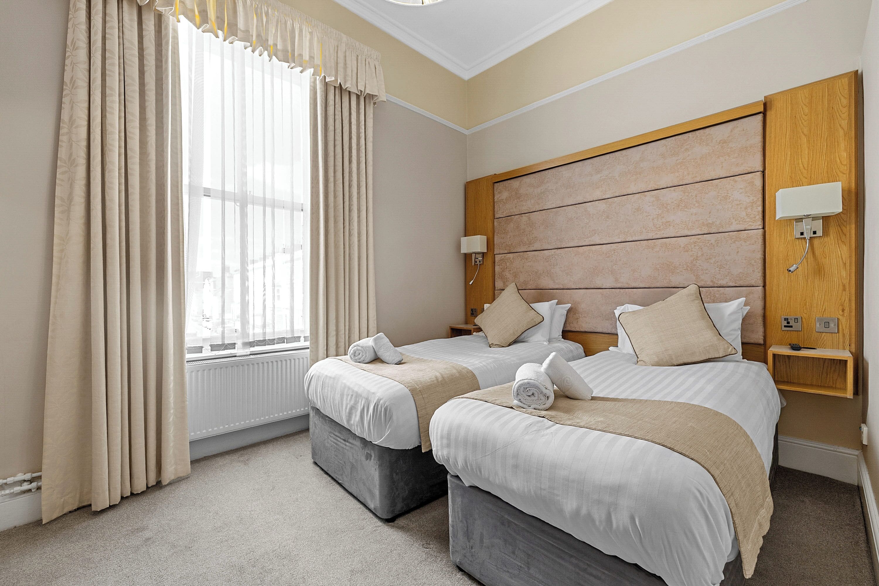 A hotel room with two beds, a large window with curtains, a wooden headboard, bedside lamps, and a small wooden shelf.