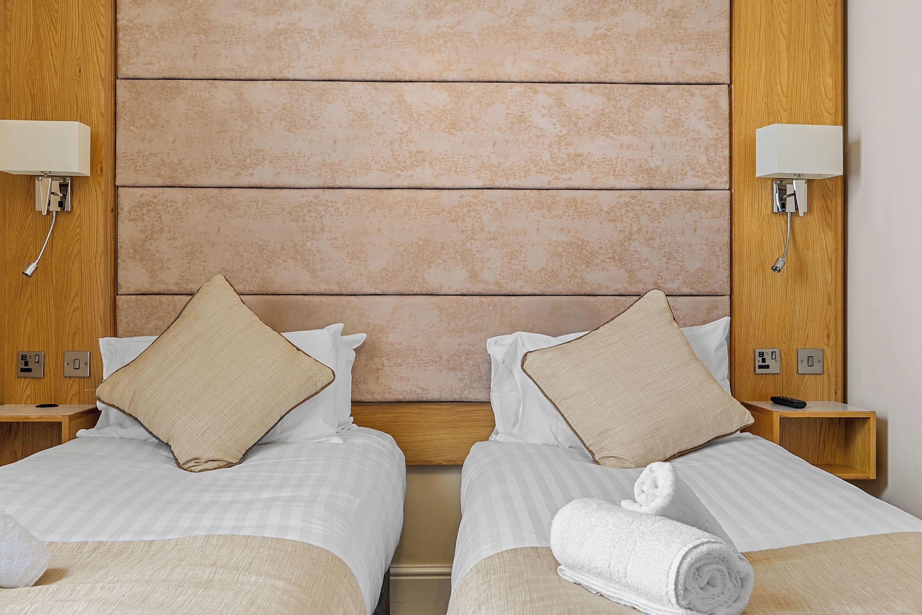 Camera d'albergo con due letti, ognuno dotato di una testiera in legno, lenzuola bianche e cuscini color beige. Sono presenti comodini con lampade e un telecomando su uno di essi.