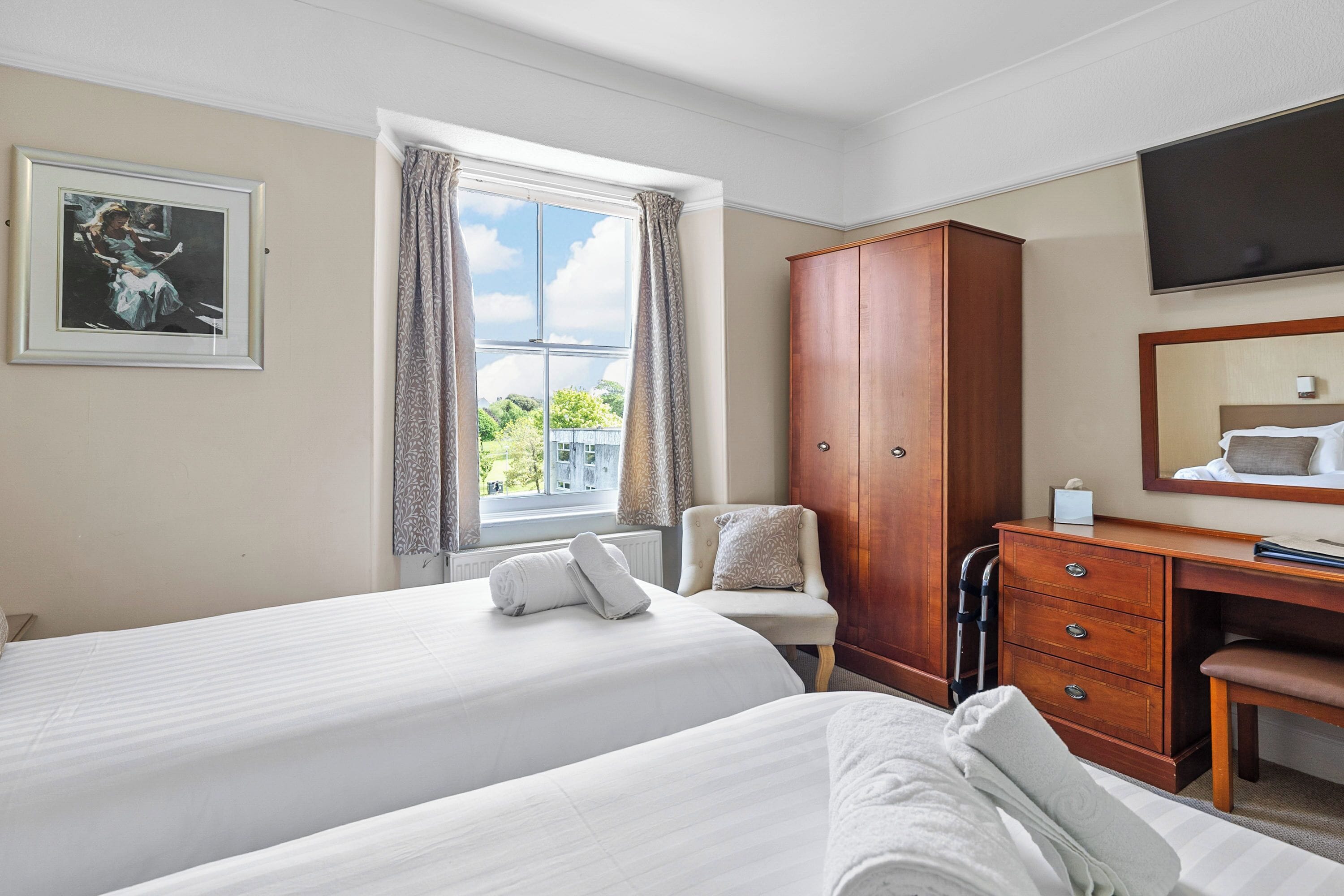 A hotel room with two beds, a wooden wardrobe, a desk, and a large window with curtains.