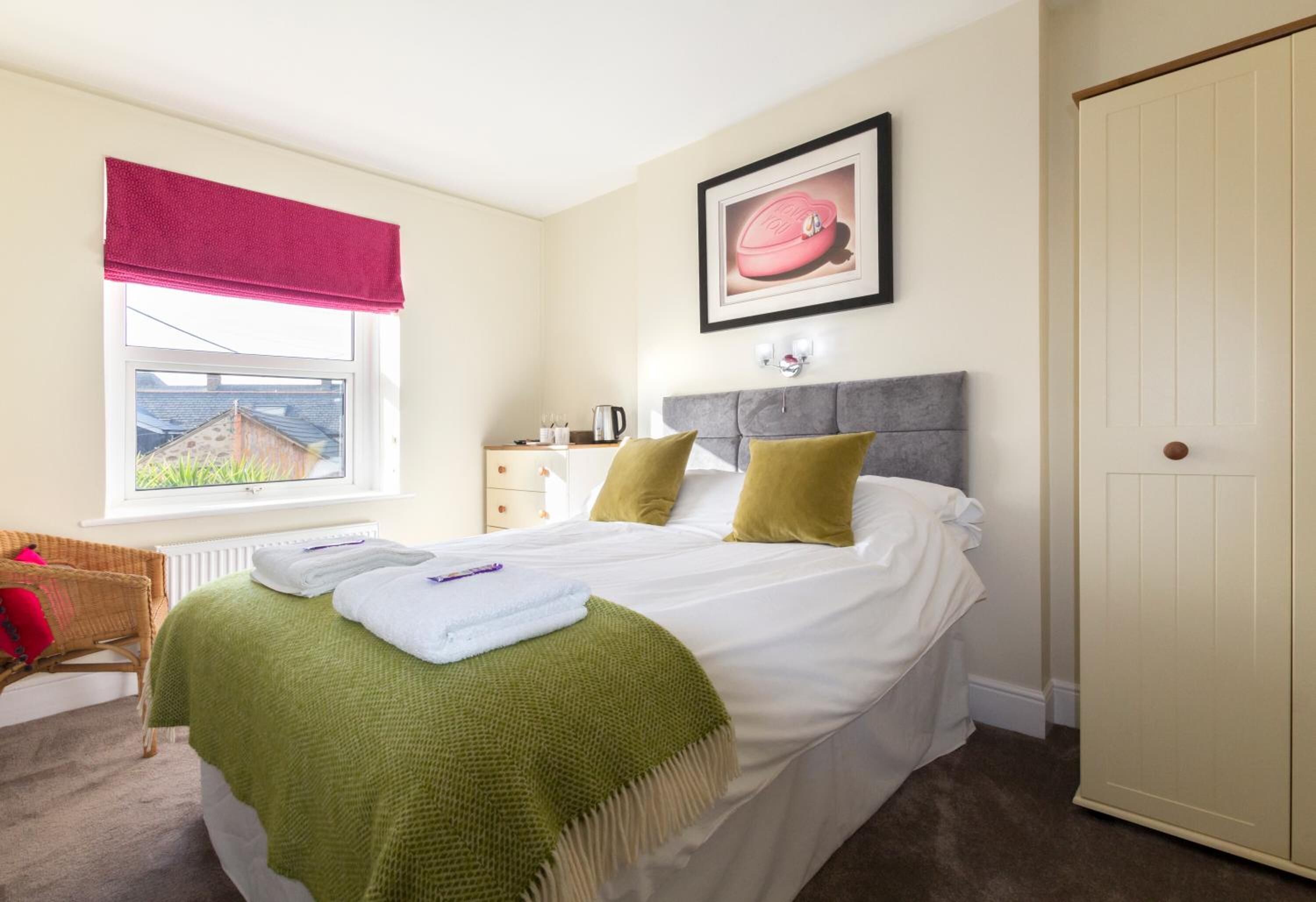A neatly made bed with a green blanket, a grey headboard, and two pillows. There is a window with a red blind, a framed picture on the wall, and a white door.