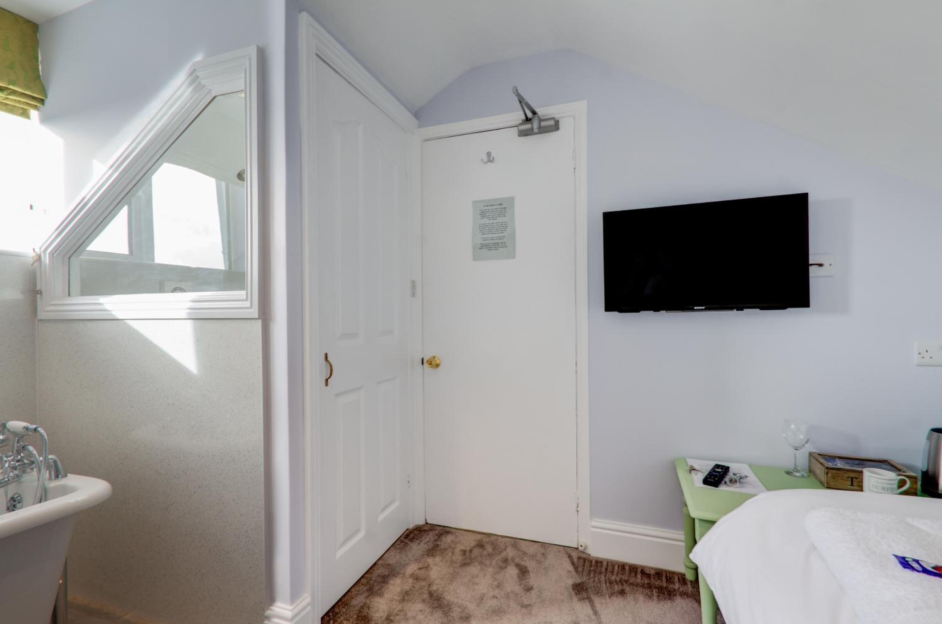 A bedroom with a white door, a mounted television, a green nightstand, and a bed with a white comforter.