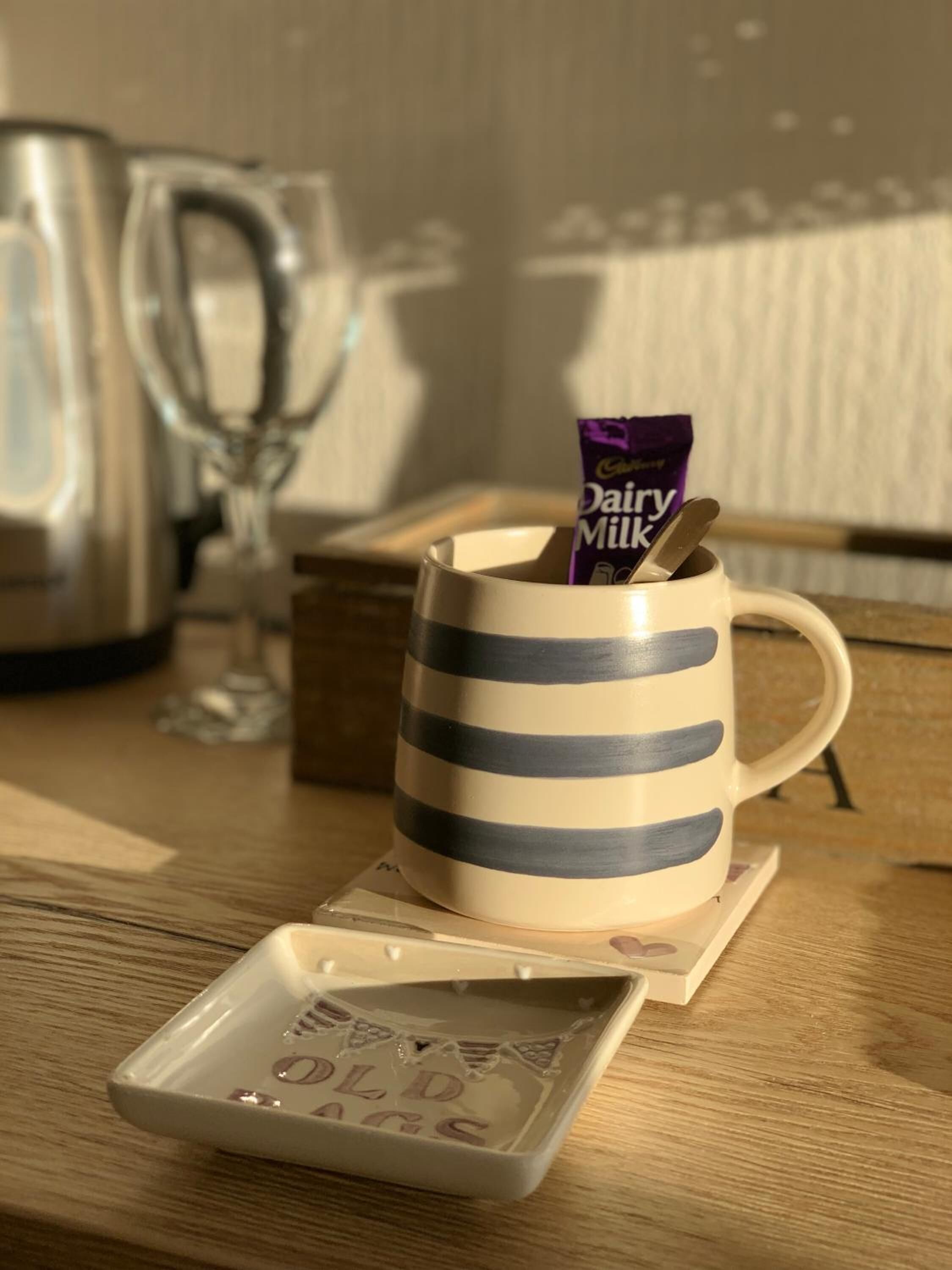 A striped mug with a chocolate bar and a spoon on a coaster, a glass, and a kettle on a wooden table.