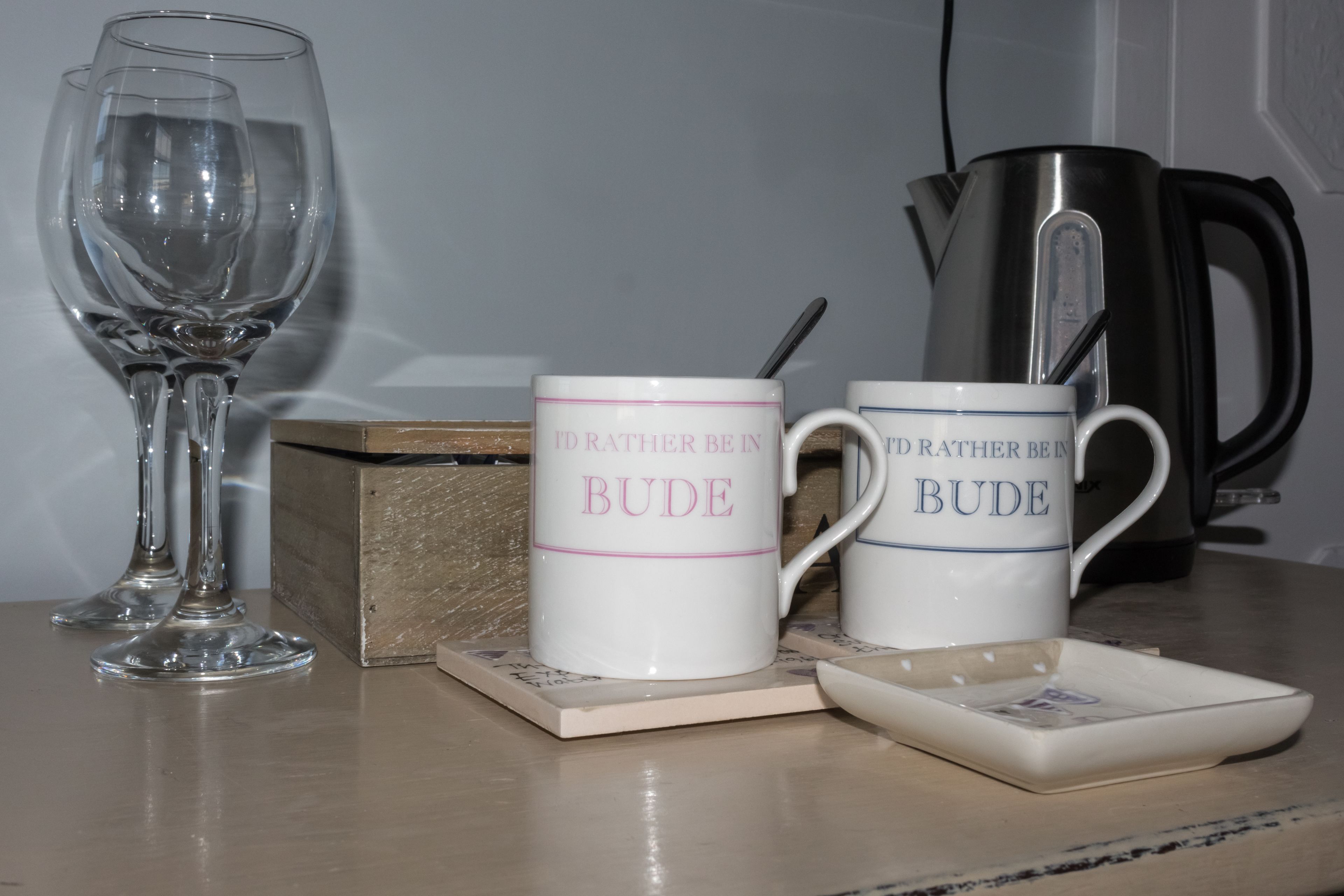 Dos tazas blancas con la inscripción "I'd rather be in Bude" sobre una mesa con un vaso y una cafetera.