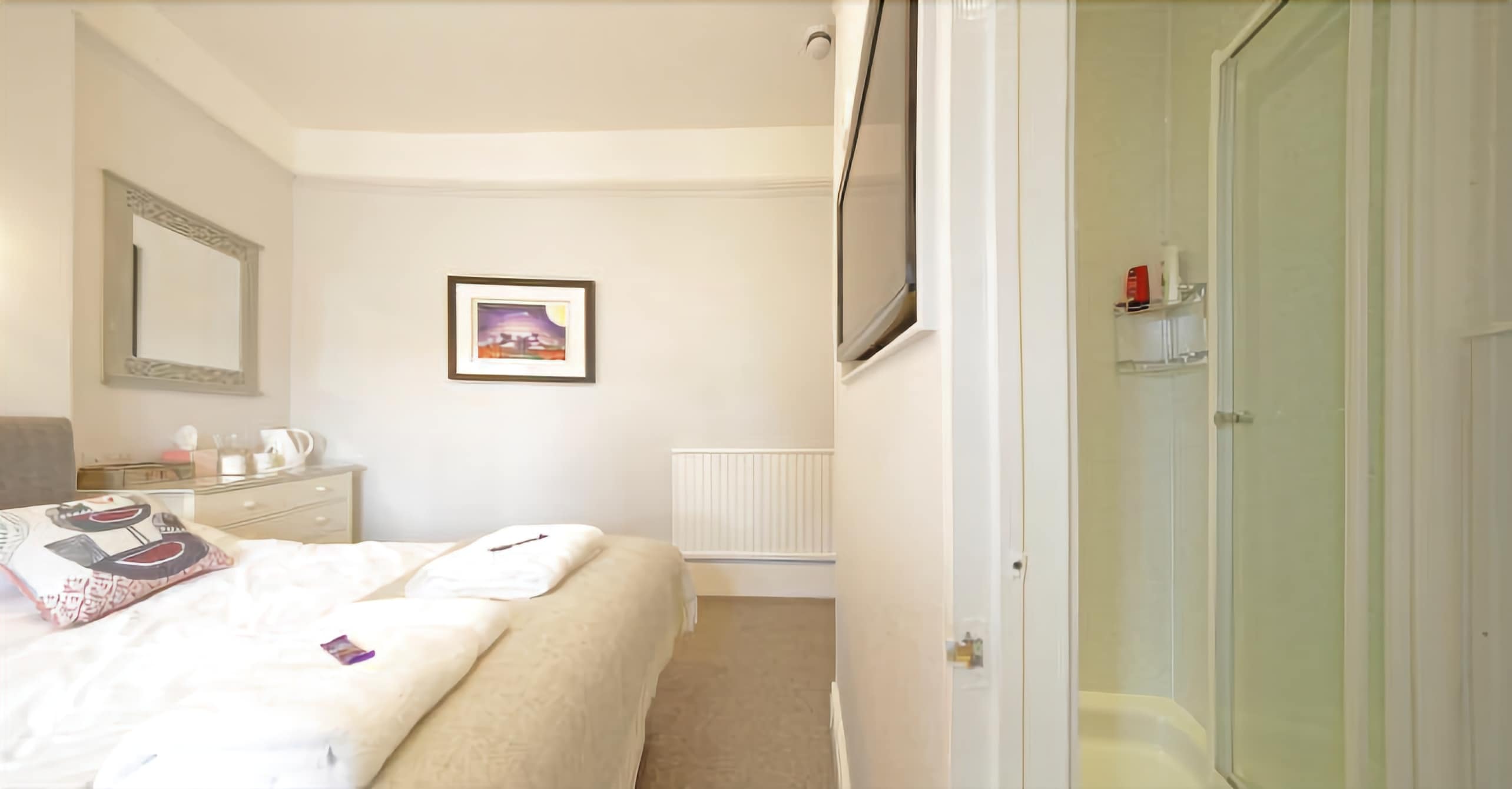A bedroom with a bed, a bedside table, a mirror, a framed picture, and a bathroom with a glass door.