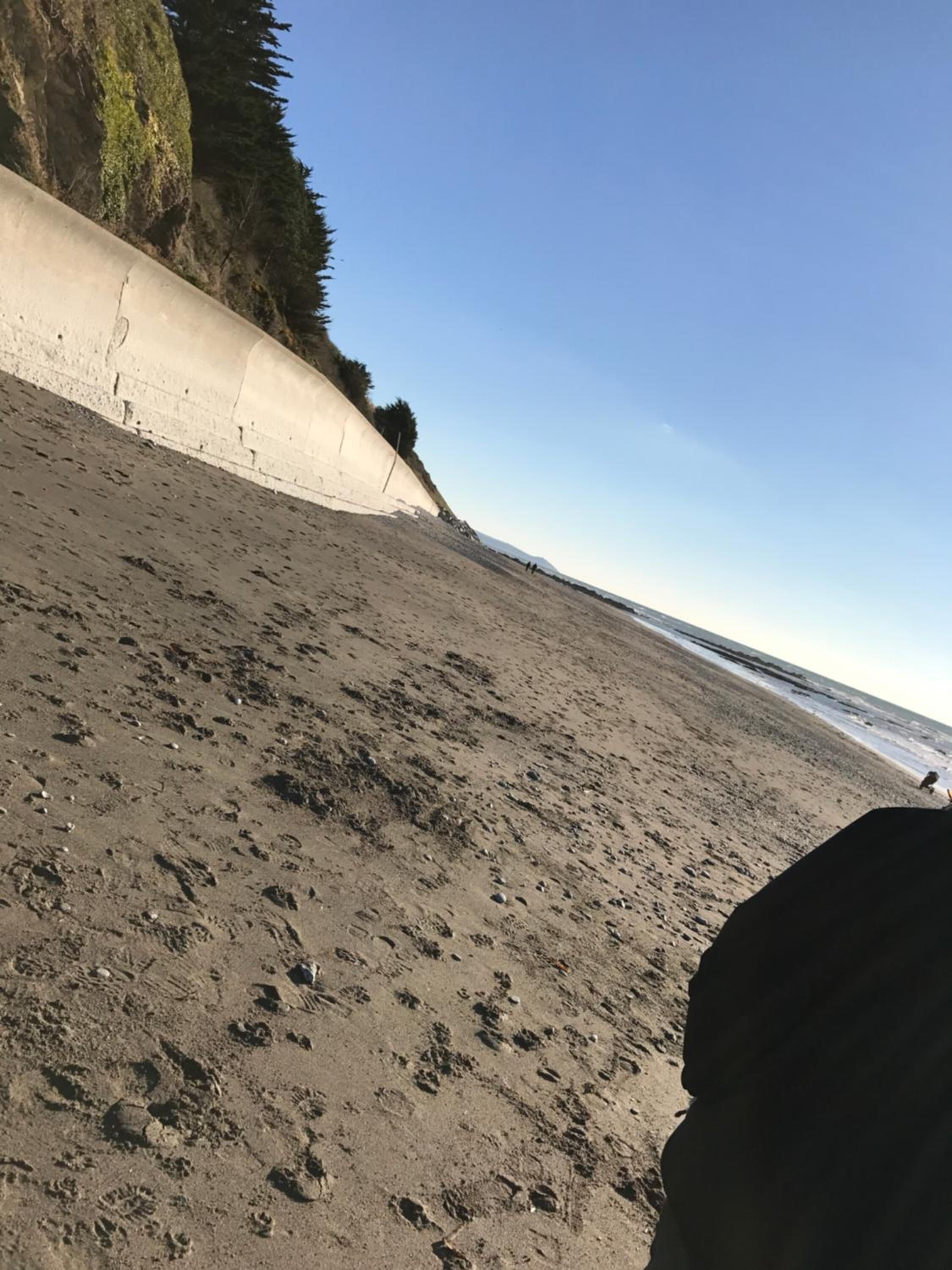 Una playa arenosa con huellas, un muro de concreto y un cielo despejado.
