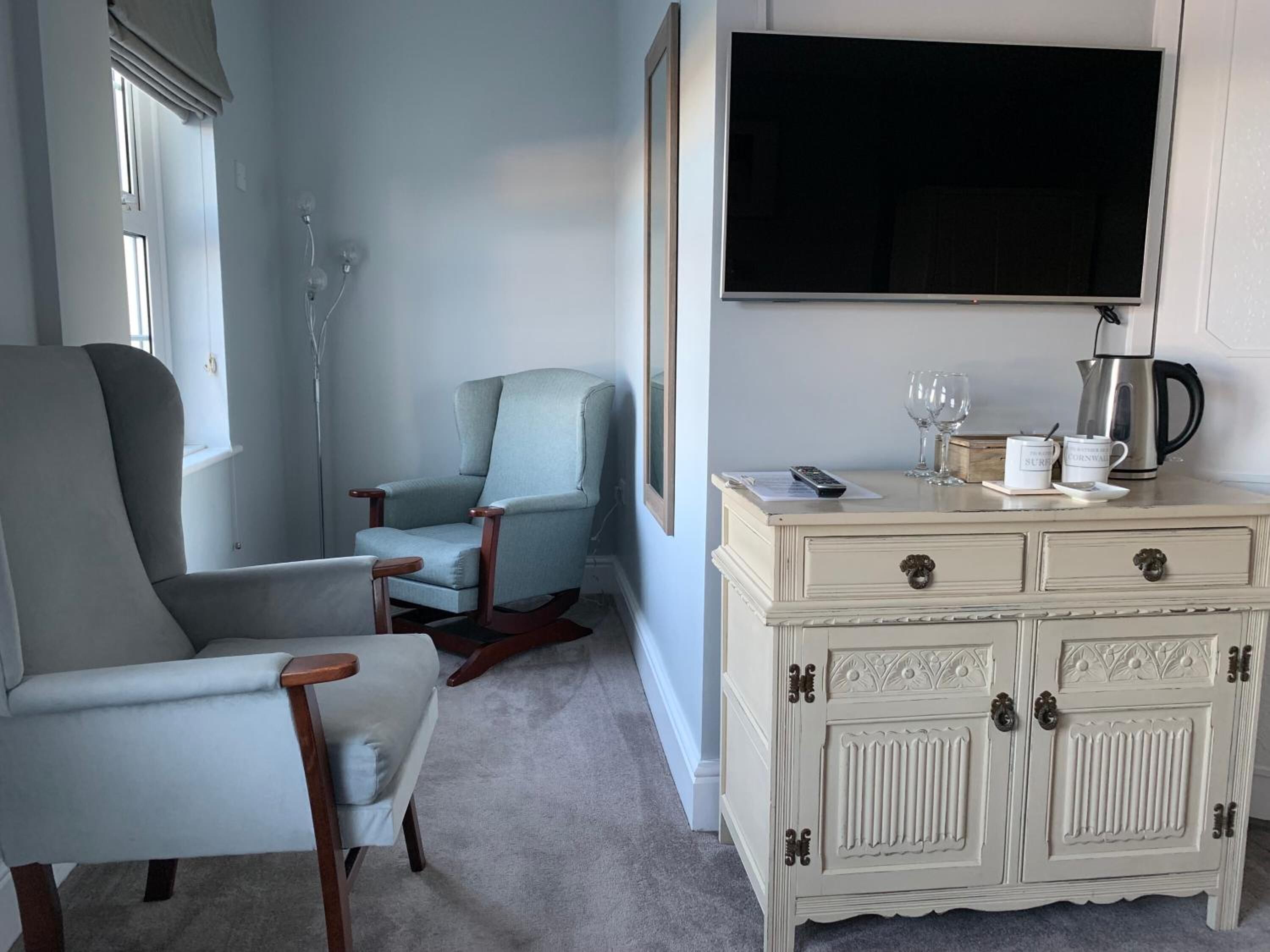 A room with a grey armchair, a blue armchair, a white cabinet with drawers, a mounted television, and a window with a curtain.