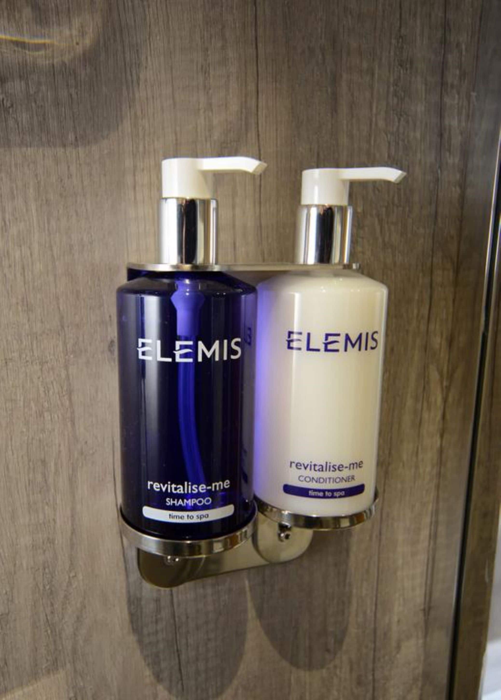 Dos productos Elemis, una botella de champú azul y una botella de acondicionador blanco, sobre una superficie de madera.
