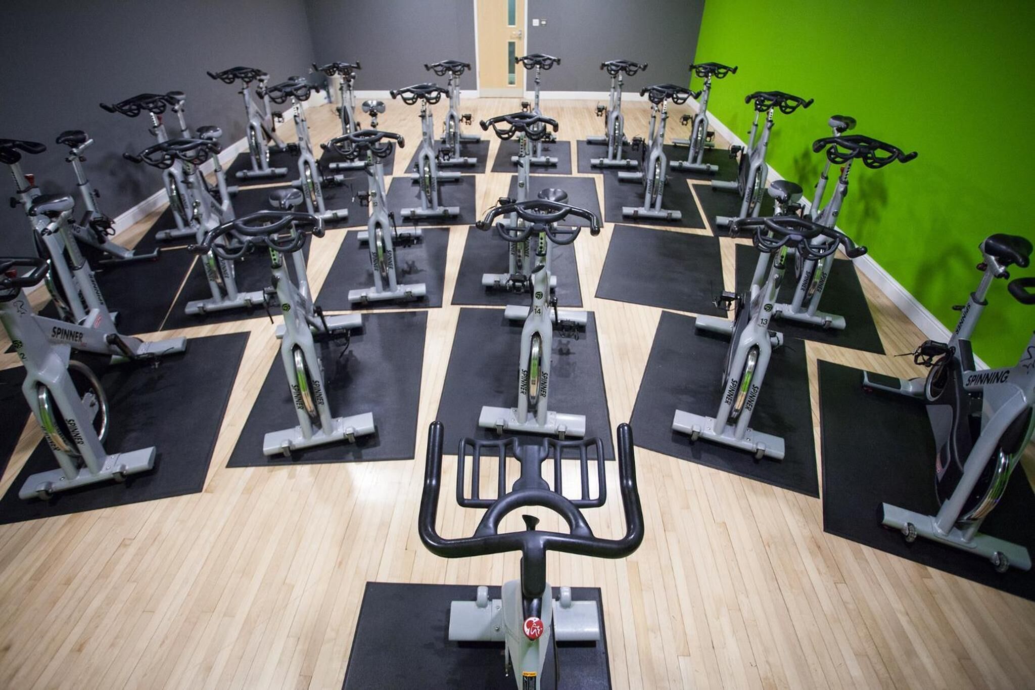 A gym with multiple stationary bikes arranged in rows on black mats, with a green wall in the background.