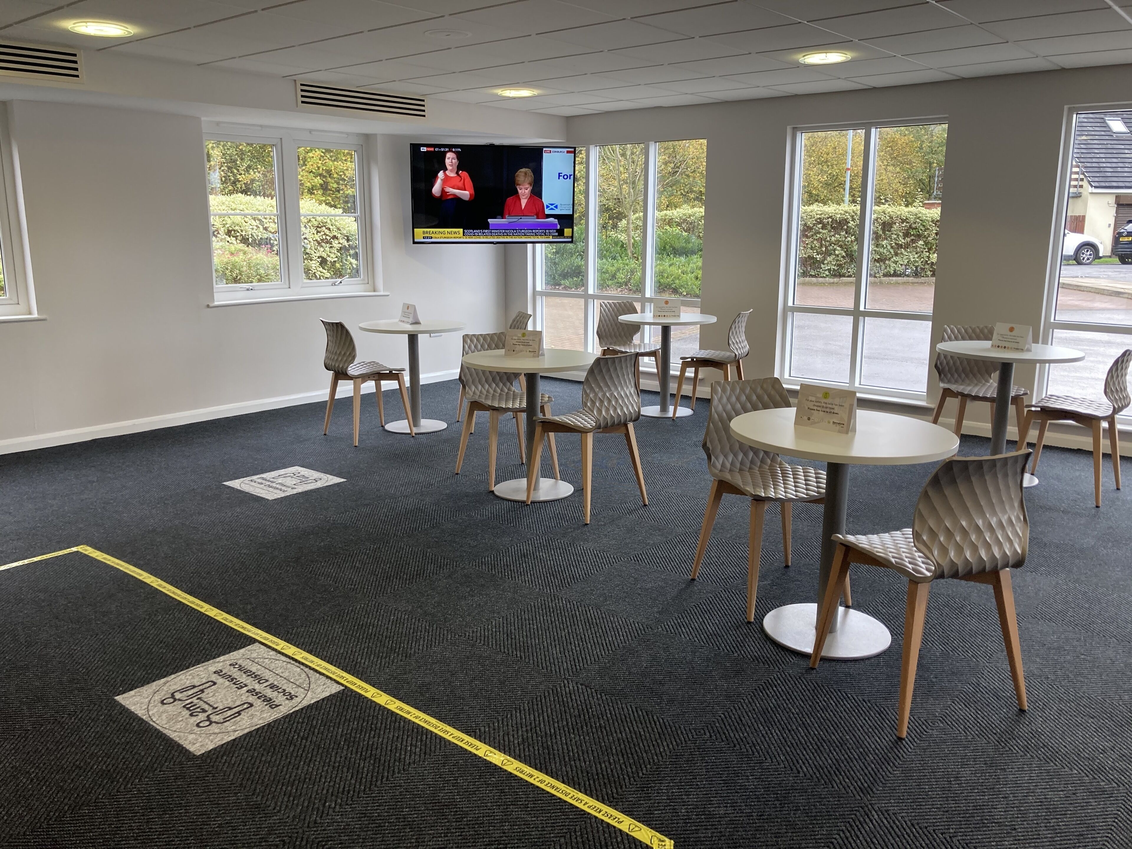 A conference room with round tables and chairs, a large screen displaying a news broadcast, and large windows with a view of the outside.