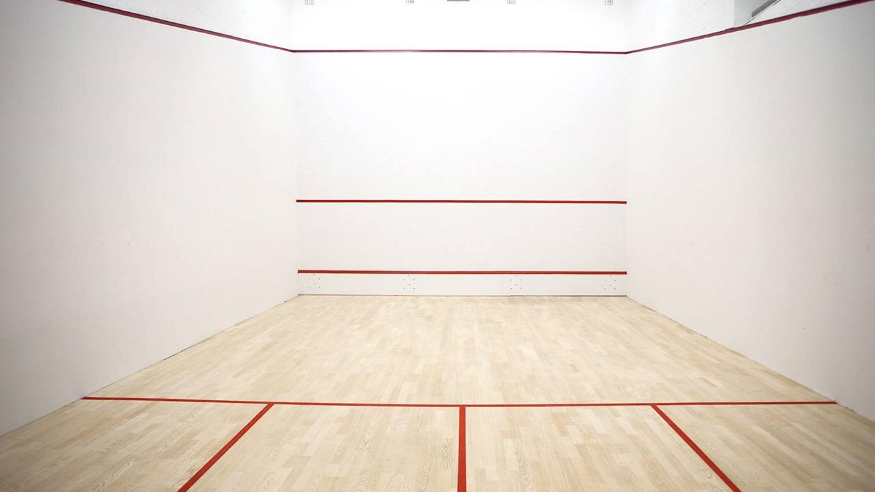 Una cancha de squash con paredes blancas y suelo de madera, marcada con líneas rojas.