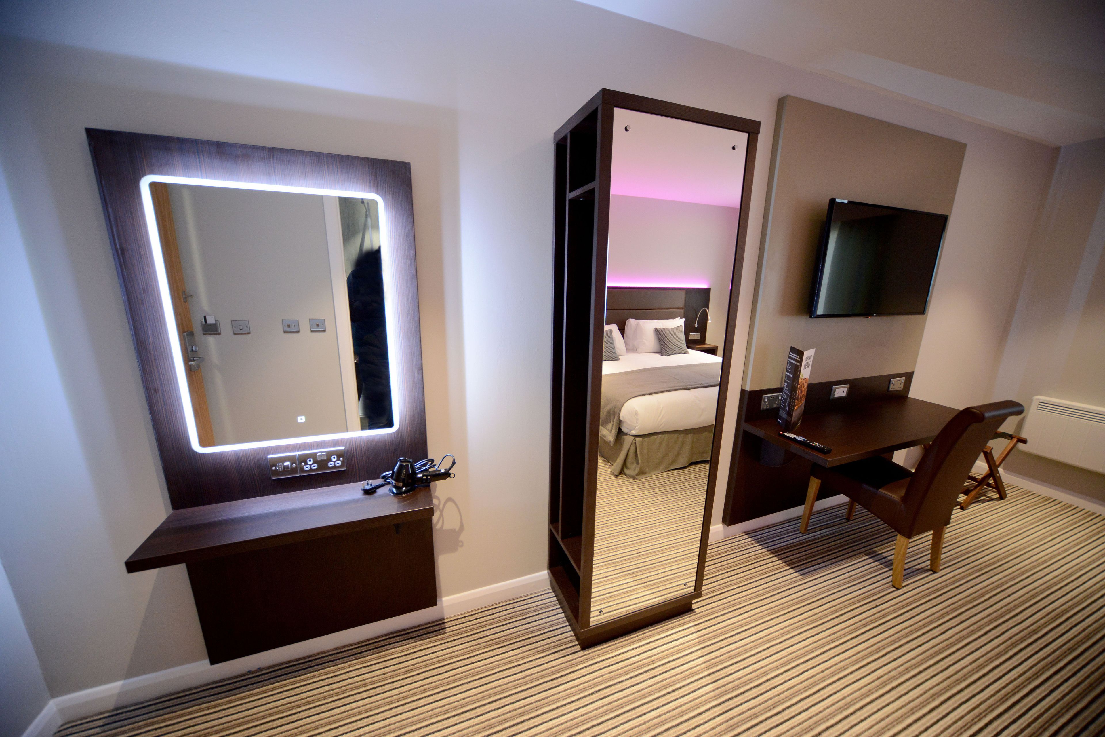 A hotel room with a large mirror, a bed, a desk, and a TV.