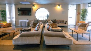 A spacious living area with a large round window, two sofas with cushions, a coffee table, and a flat-screen TV mounted on the wall. - Club Vacances Bleues Les Jardins Atlantique (Talmont-Saint-Hilaire)