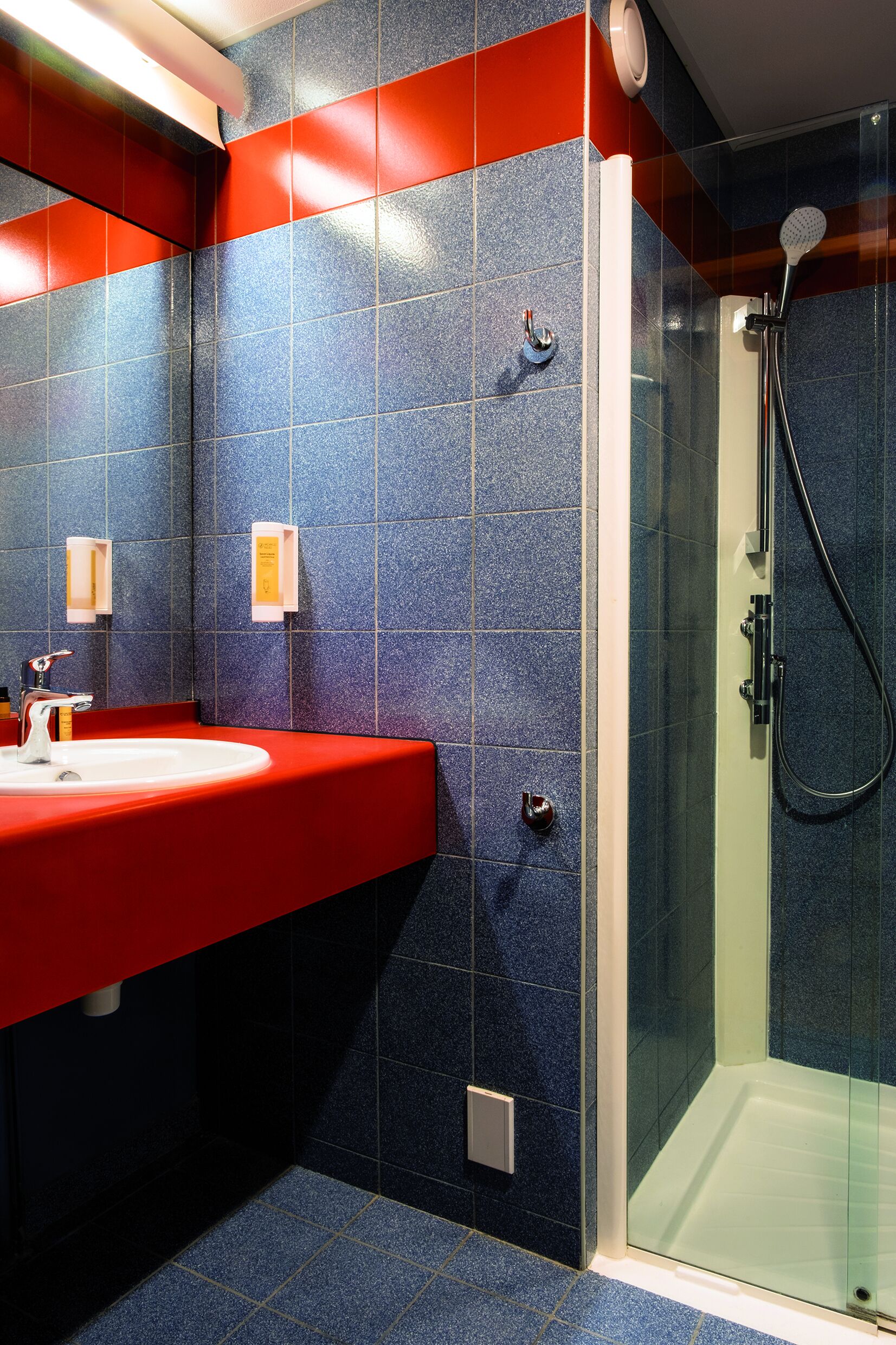 A bathroom with a red sink, blue tiles, a glass shower enclosure, and a white towel dispenser.