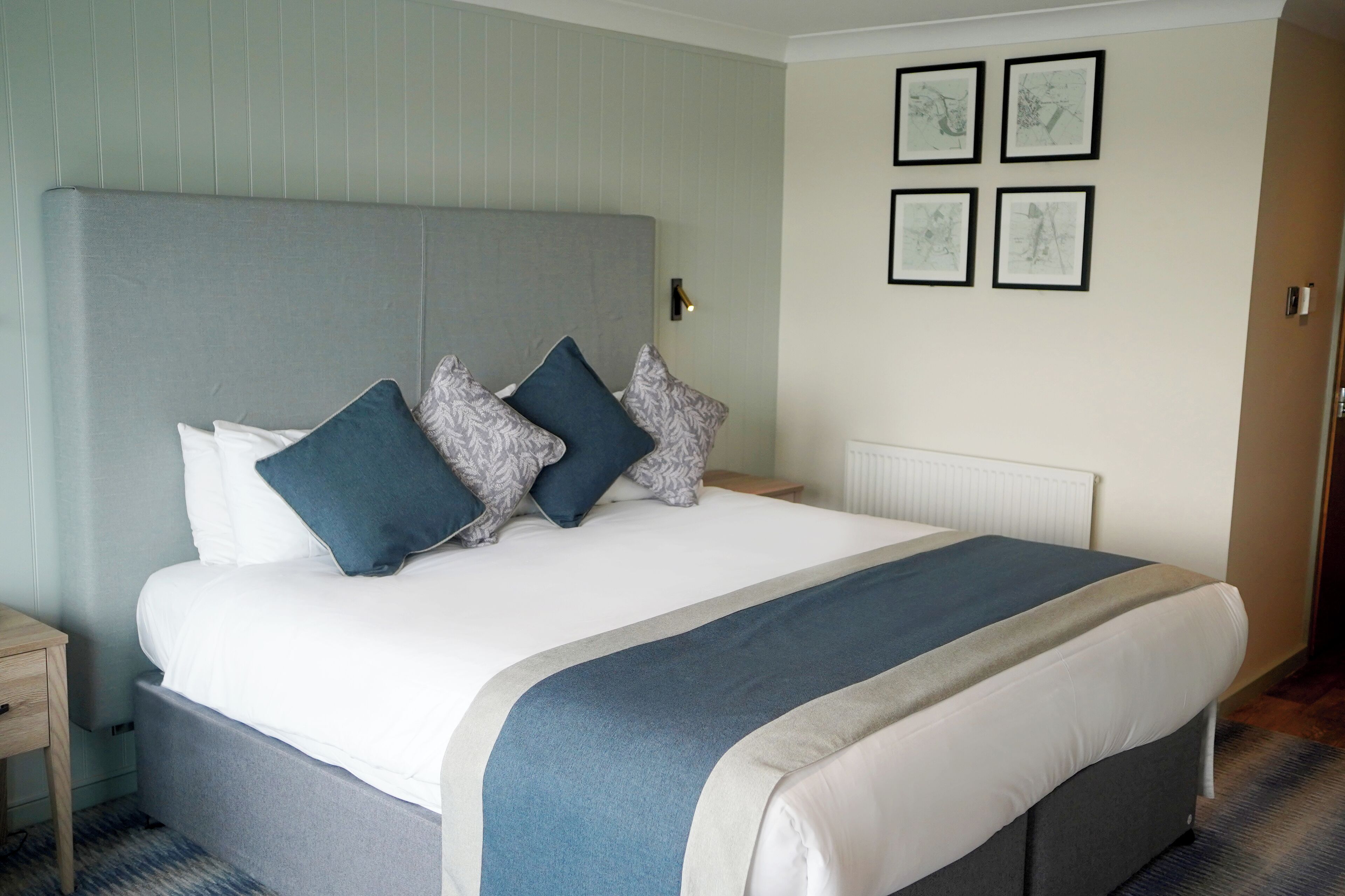 A neatly made bed with a grey headboard, white bedding, and a blue accent throw. There are four framed pictures on the wall above the bed.