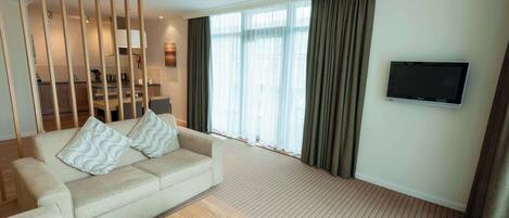 Habitación de hotel con sofá, mesa de centro, televisor y un ventanal con cortinas.