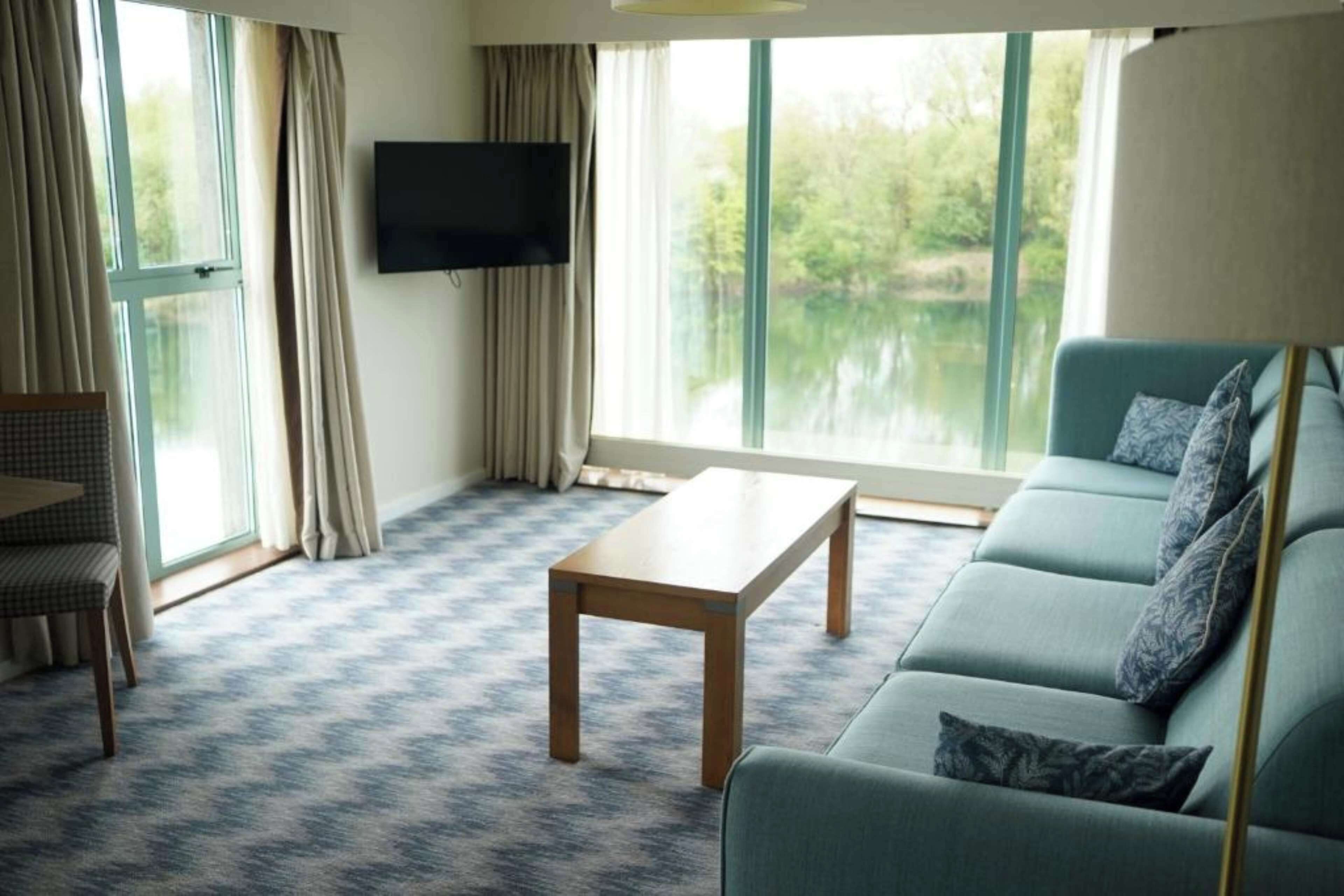 A modern living room with a sofa, a coffee table, a TV mounted on the wall, and large windows with a view of greenery.