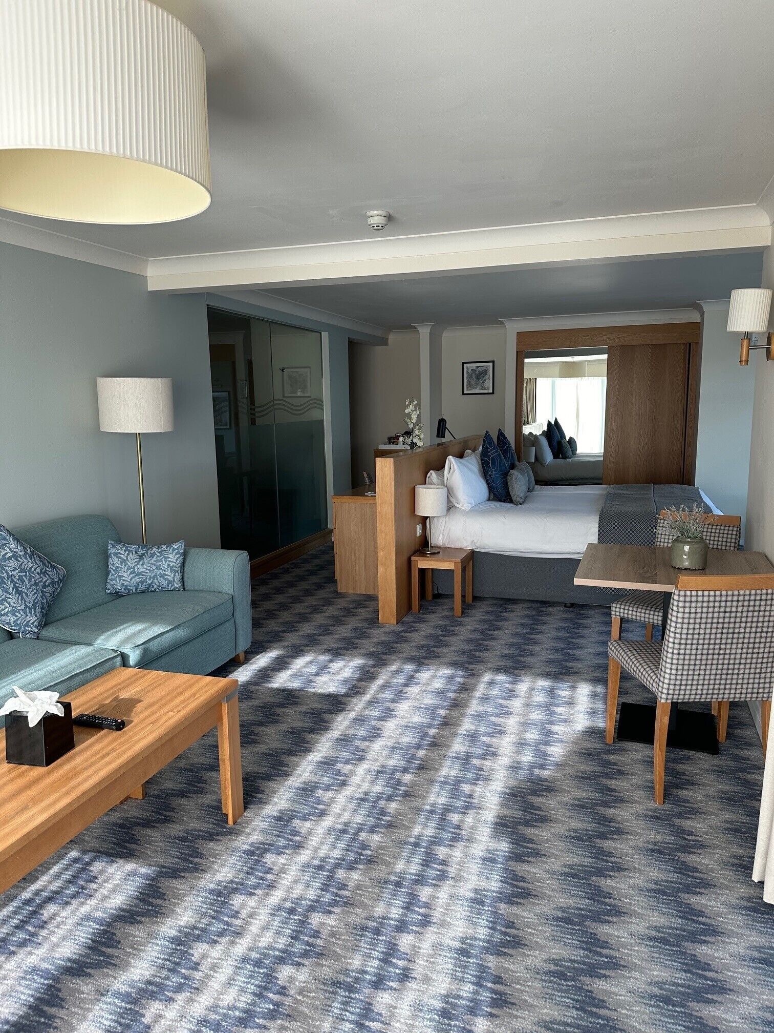 A modern hotel room with a sofa, a bed, a dining table with chairs, a lamp, and a carpeted floor.