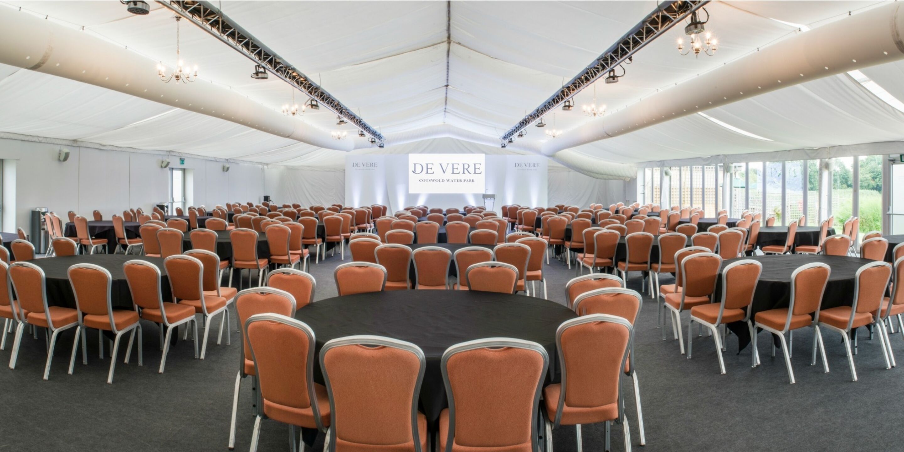 A large conference room with orange chairs and tables, a white ceiling, and a sign that reads "DEVERE COTSWOOD WATER PARK".