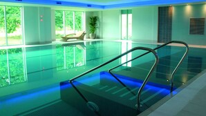 A modern indoor swimming pool with clear glass walls, a sunlit area with a lounge chair, and a potted plant. - De Vere Cotswold Water Park (Cirencester)