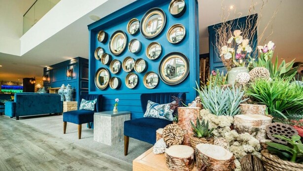 A modern lobby with a blue accent wall, multiple mirrors, and a variety of decorative plants and wooden elements. - De Vere Cotswold Water Park (Cirencester)