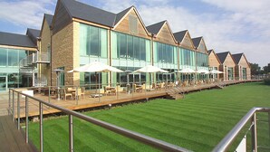 A modern hotel with a long outdoor deck, large windows, and outdoor seating with umbrellas. - De Vere Cotswold Water Park (Cirencester)