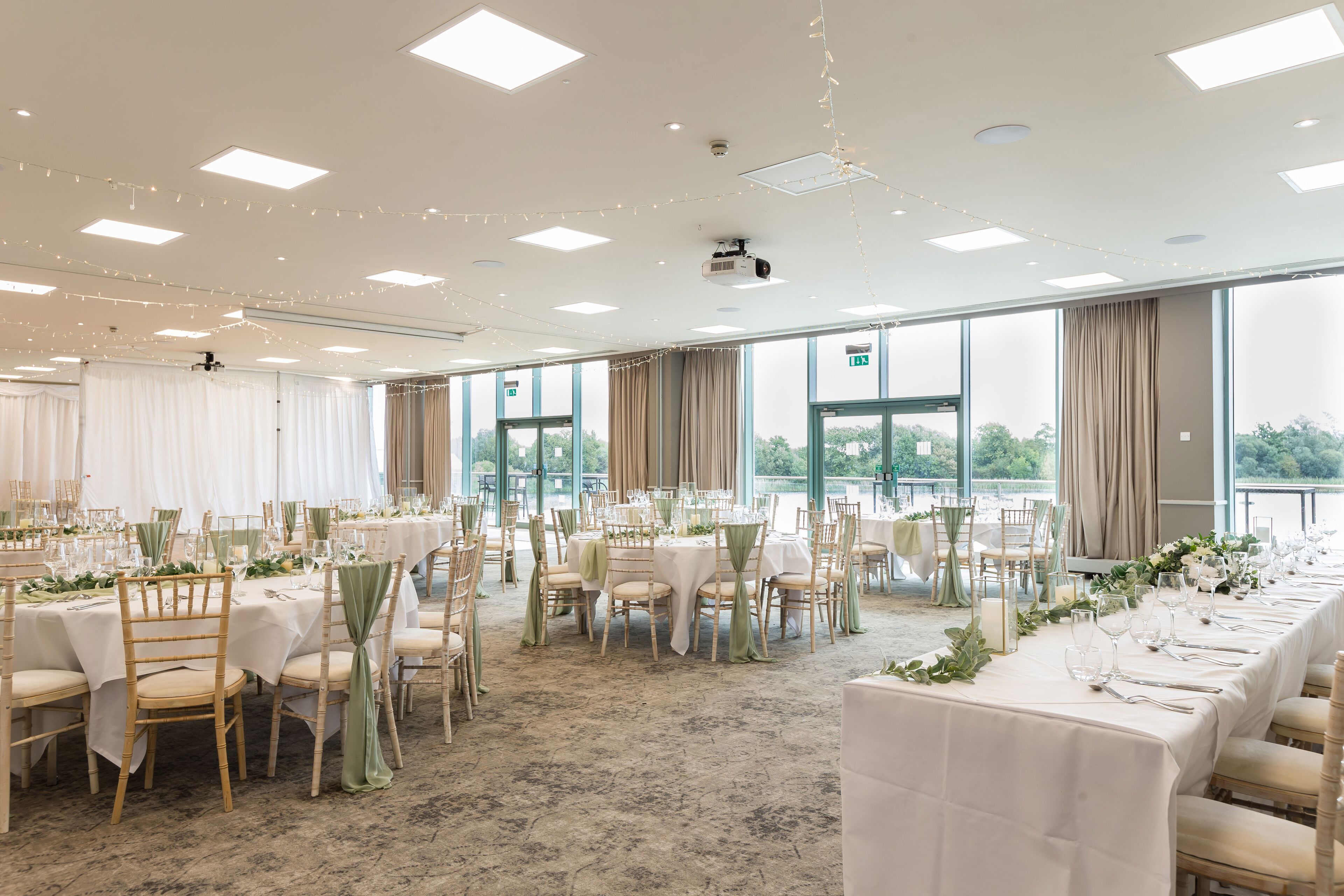 A banquet hall with multiple round tables, each set with chairs and tablecloths, and large windows offering a view of the outdoors.
