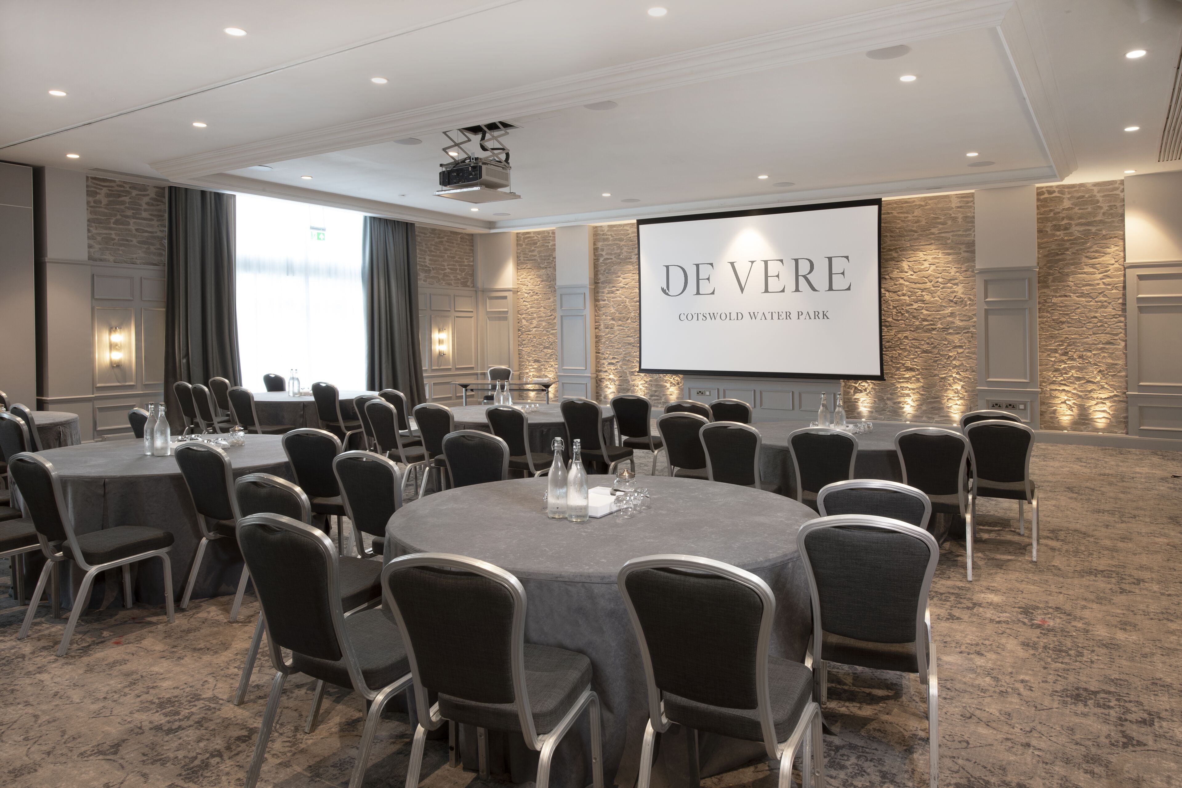 A conference room with round tables and chairs, a projector screen, and a stone wall.