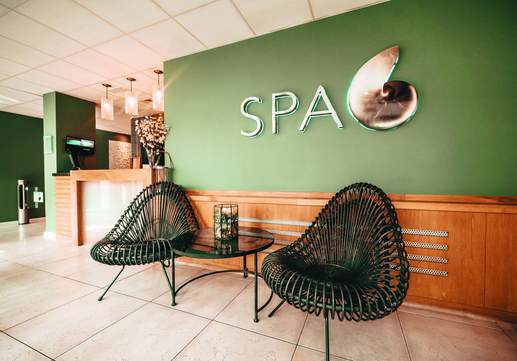 Zona de recepción con pared verde, un letrero que dice 'SPA', dos sillones de mimbre negro, una mesita y un mostrador de recepción con un monitor.