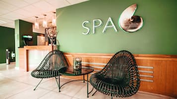 Zona de recepción con pared verde, un letrero que dice 'SPA', dos sillones de mimbre negro, una mesita y un mostrador de recepción con un monitor.
