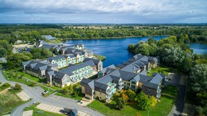 A residential area with modern houses by a river, featuring a central communal building and a parking area. - De Vere Cotswold Water Park (Cirencester)