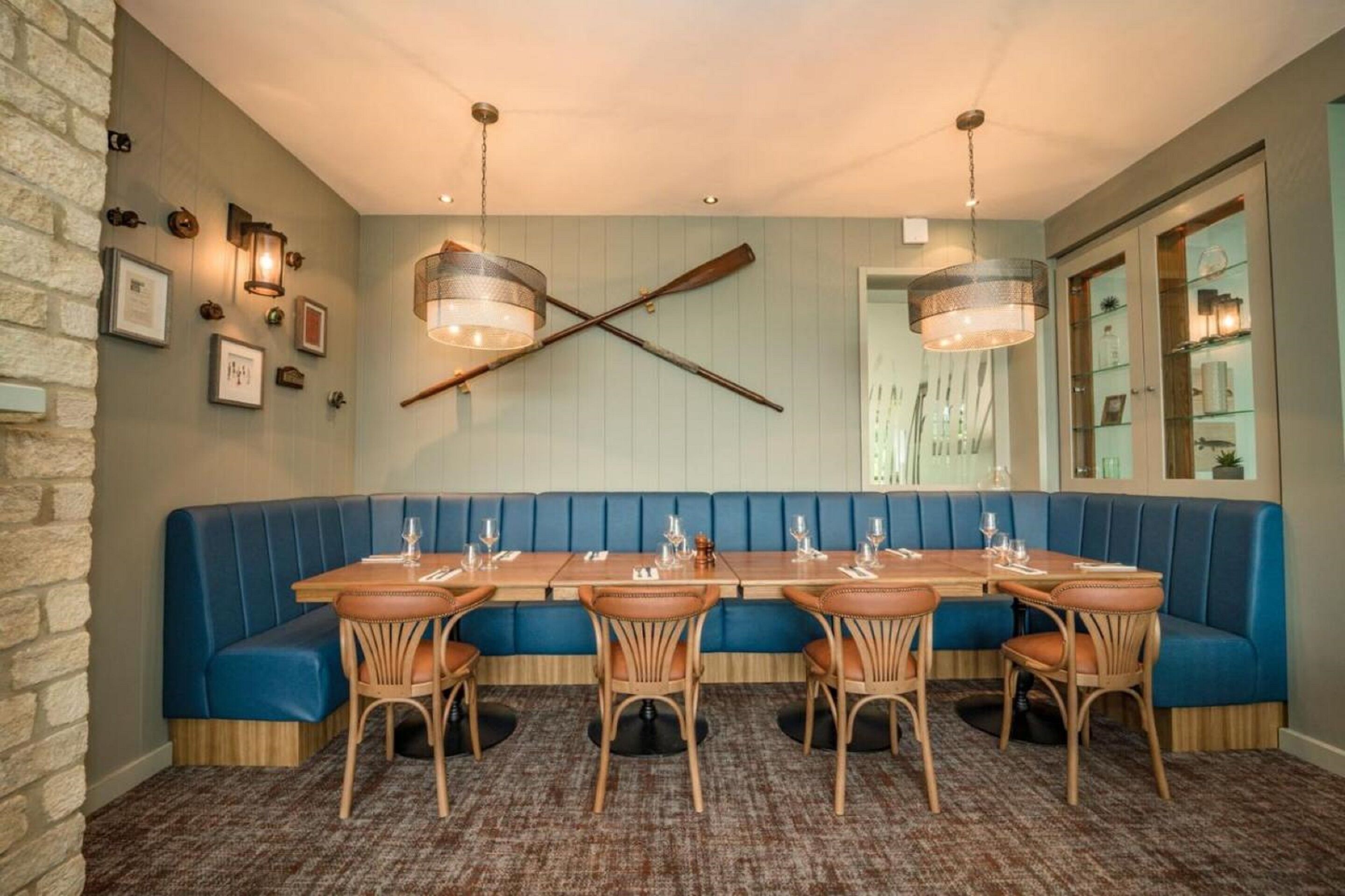 Um restaurante com um amplo banco azul, mesas e cadeiras de madeira. Há luminárias na parede e uma grande janela com vista.