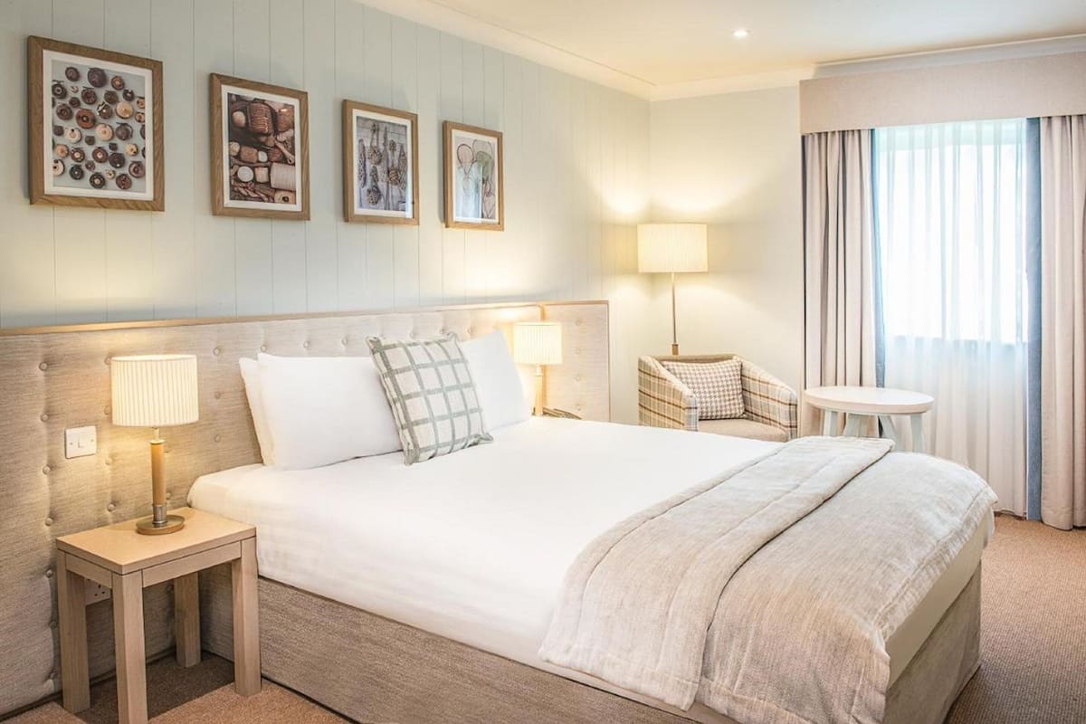 Ein Hotelzimmer mit einem großen Bett, zwei Nachttischen mit Lampen, einem Sessel, einem kleinen runden Tisch und einem Fenster mit Vorhängen.
