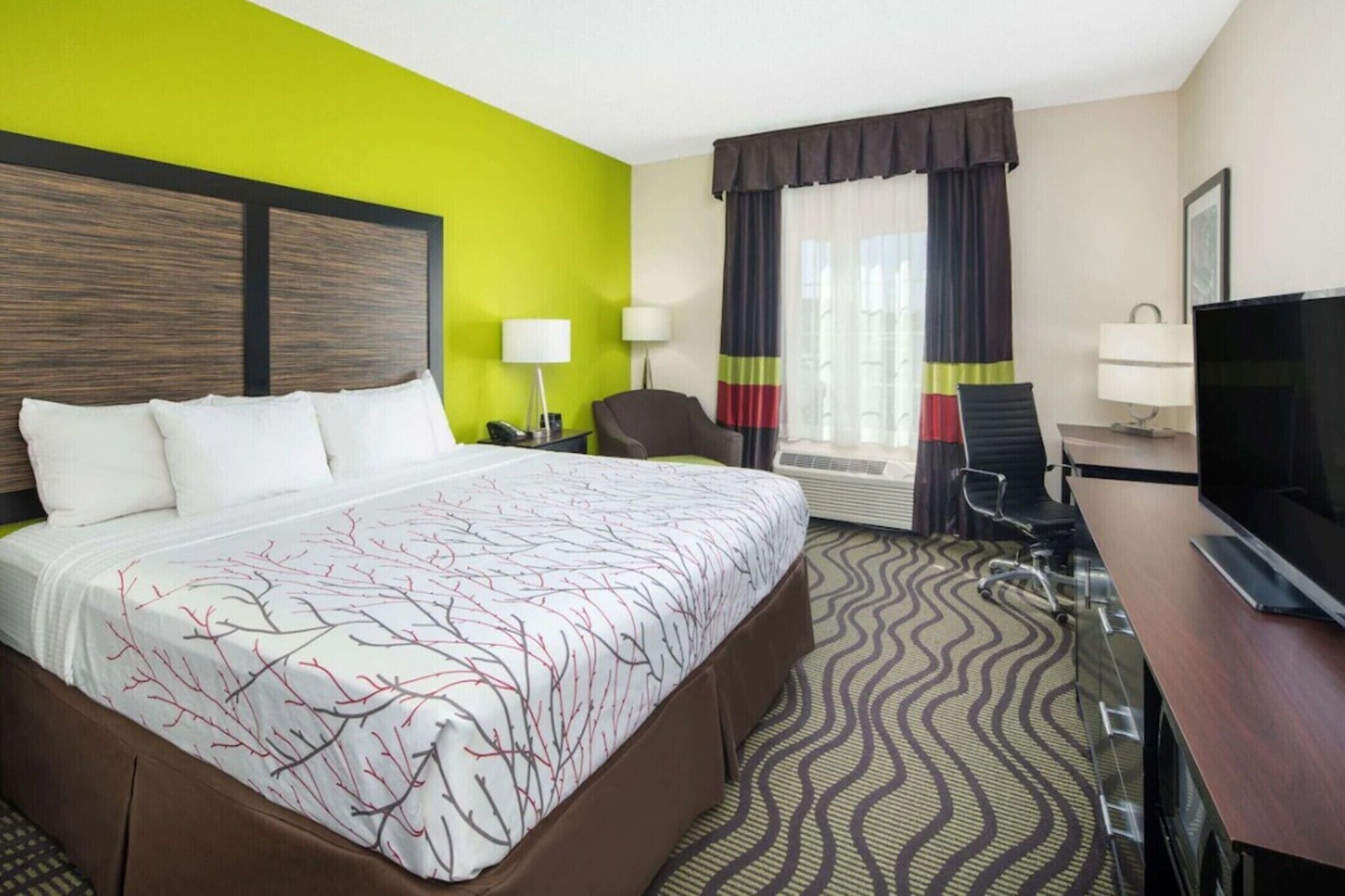 Habitación de hotel con cama, escritorio, silla, televisión y ventana con cortinas.