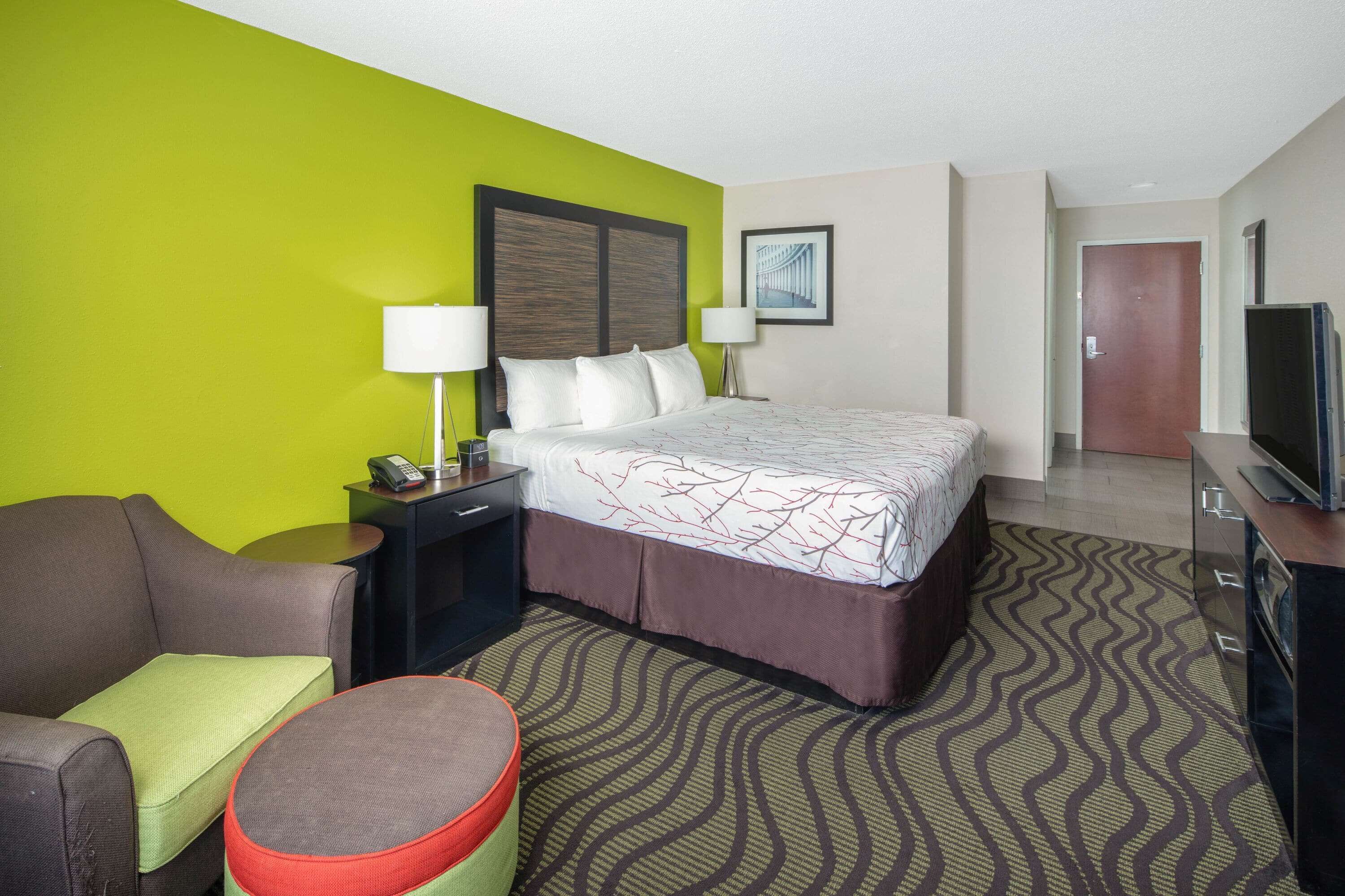 A hotel room with a bed, a desk with a lamp, a television, and a chair.
